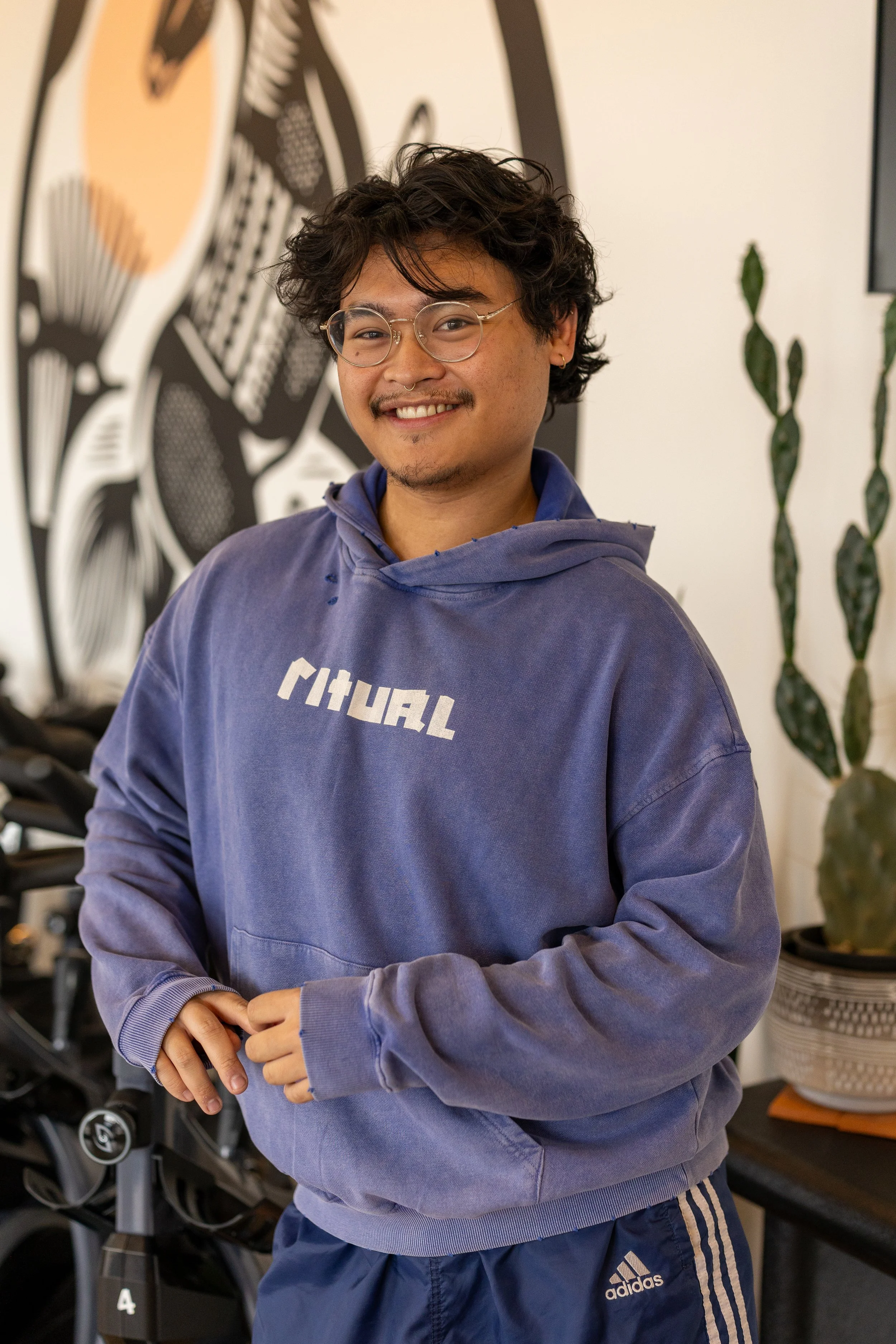 A young man with glasses smiling, wearing a purple hoodie and blue Adidas pants, standing in a gym with large exercise machines and plants in the background.