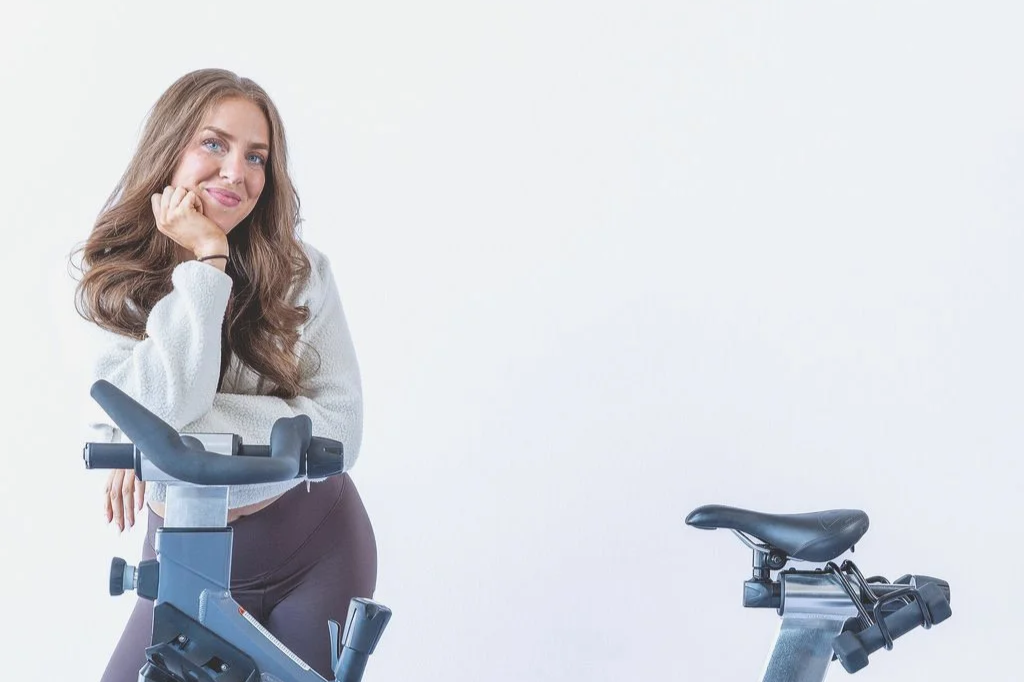 Woman with long brown hair smiling, leaning on exercise bike handlebar, in light-colored sweater and dark pants, against a plain white background.