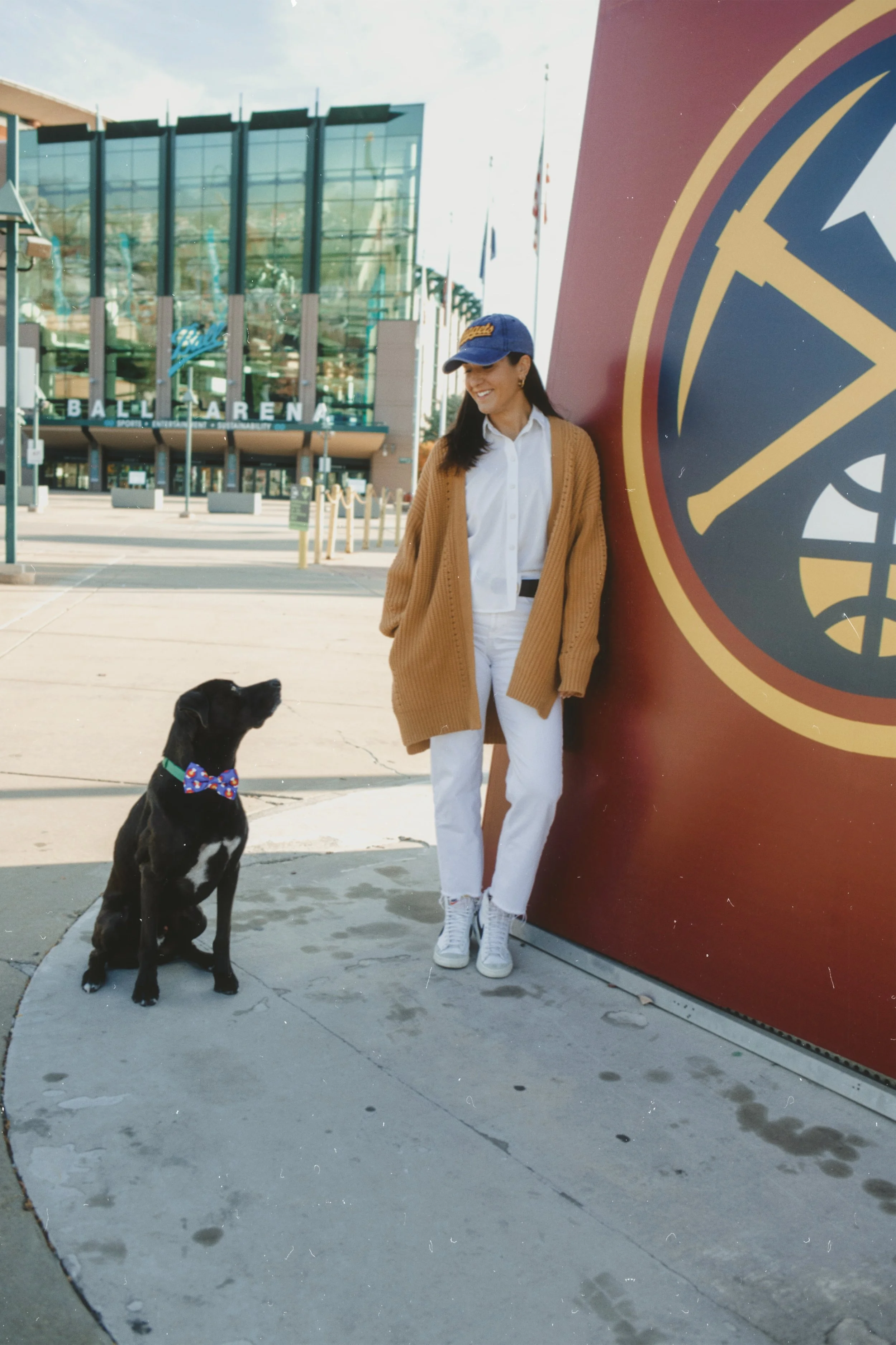 A person wearing a blue cap, white shirt, and brown cardigan stands next to a large emblem in front of Ball Arena. A black dog with a colorful bow tie sits nearby.