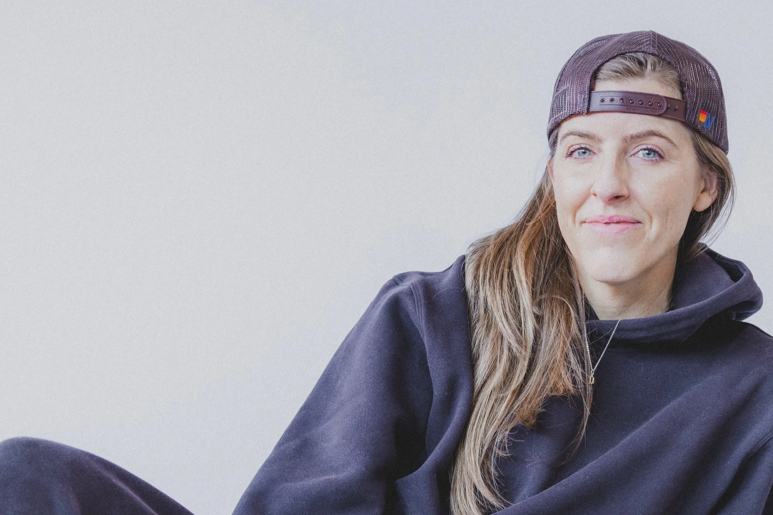 A woman with long blonde hair wearing a purple baseball cap backwards and a dark hoodie, looking at the camera with a slight smile, sitting against a plain light gray background.
