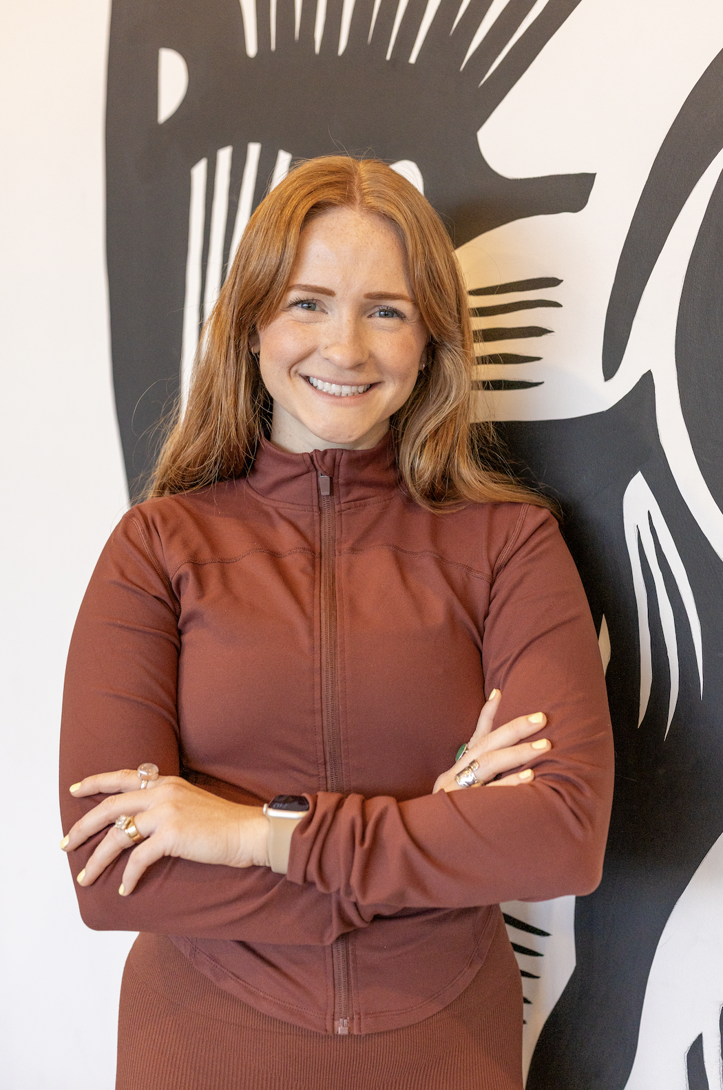 A woman with long red hair smiling, wearing a maroon zip-up jacket and standing with her arms crossed in front of a black-and-white abstract wall art.