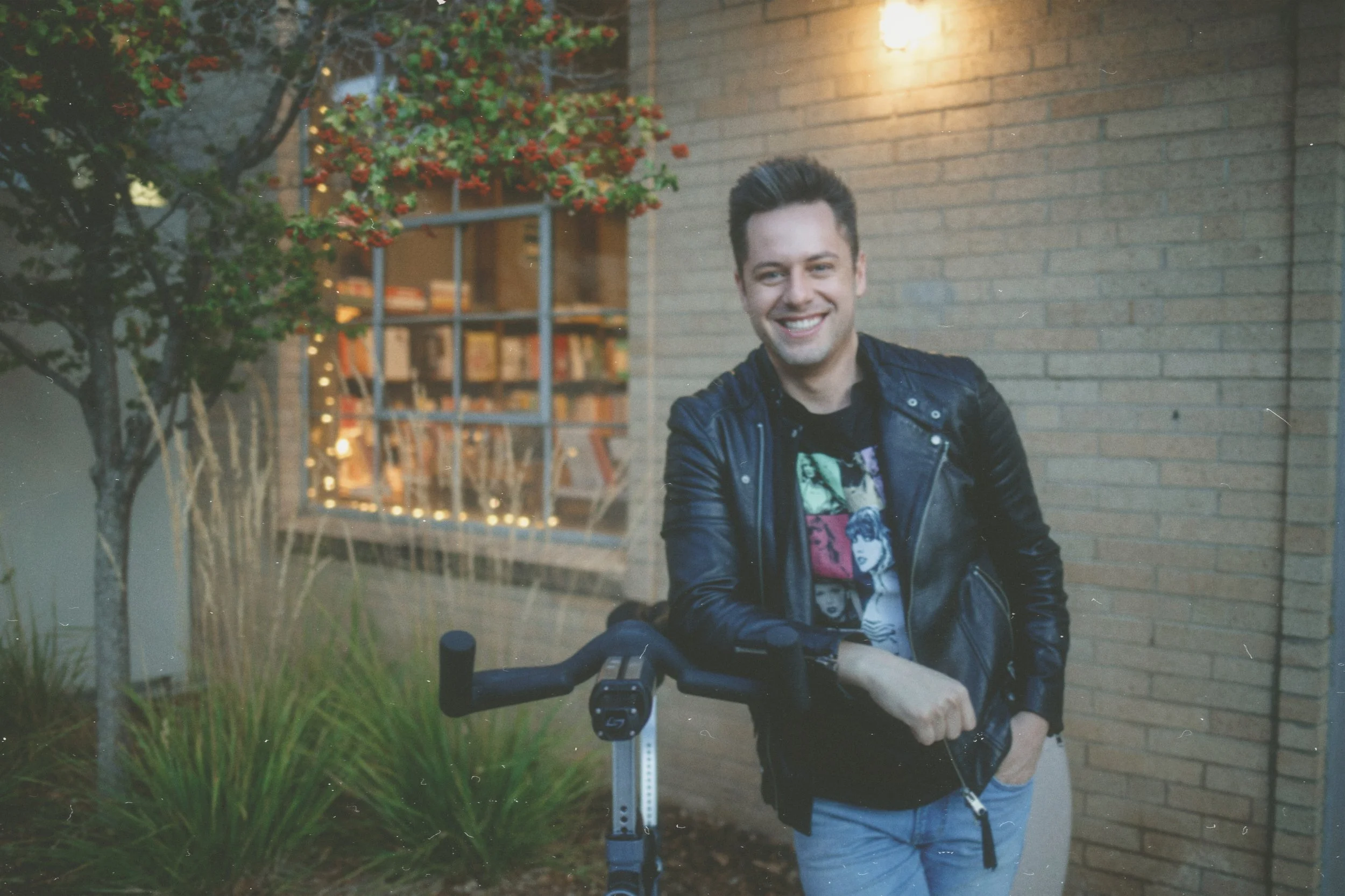 Smiling person in a leather jacket standing next to a bicycle outside a brick building with decorative lights and greenery.