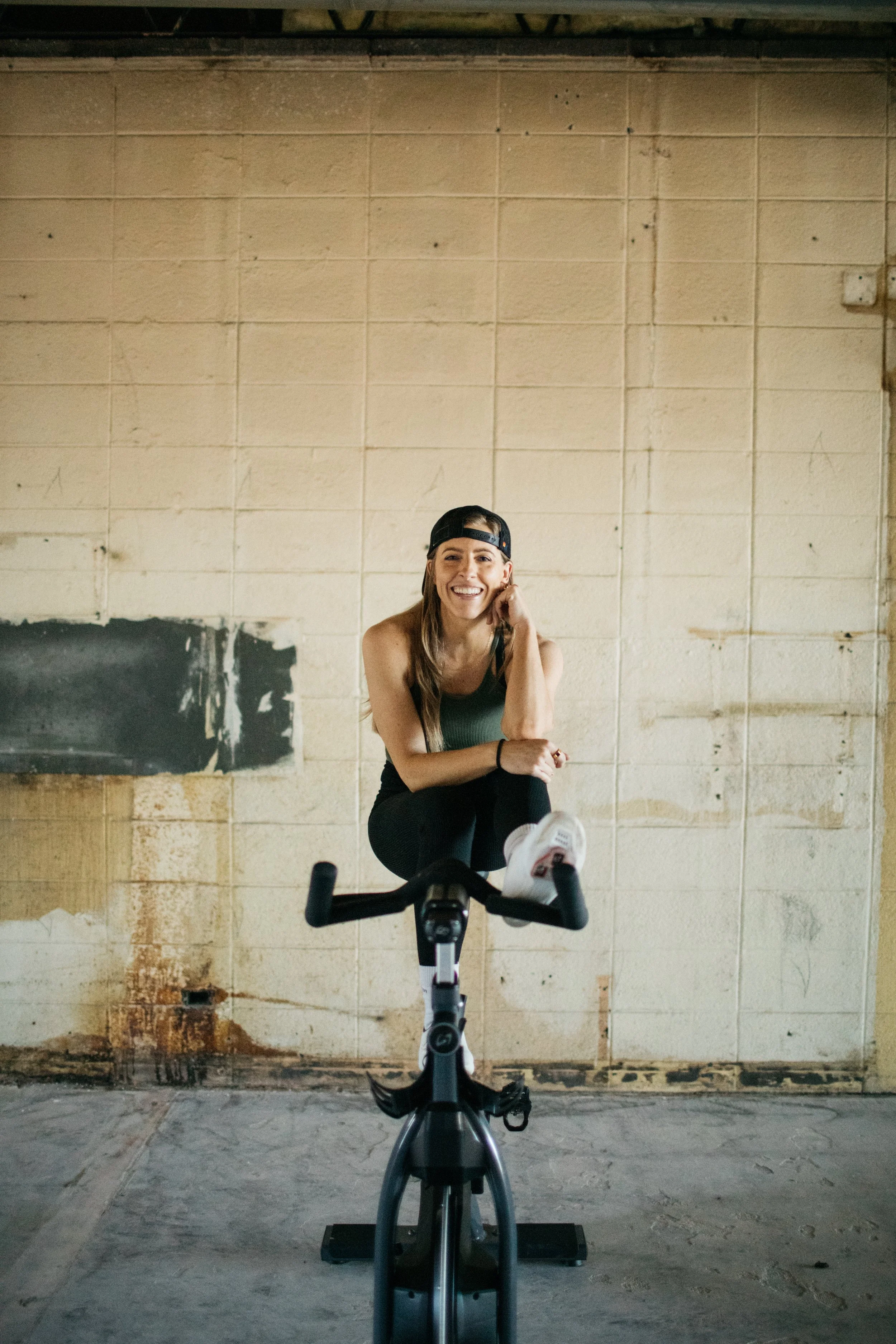 Person sitting on stationary bike in an industrial space, wearing a cap and workout clothes, smiling.