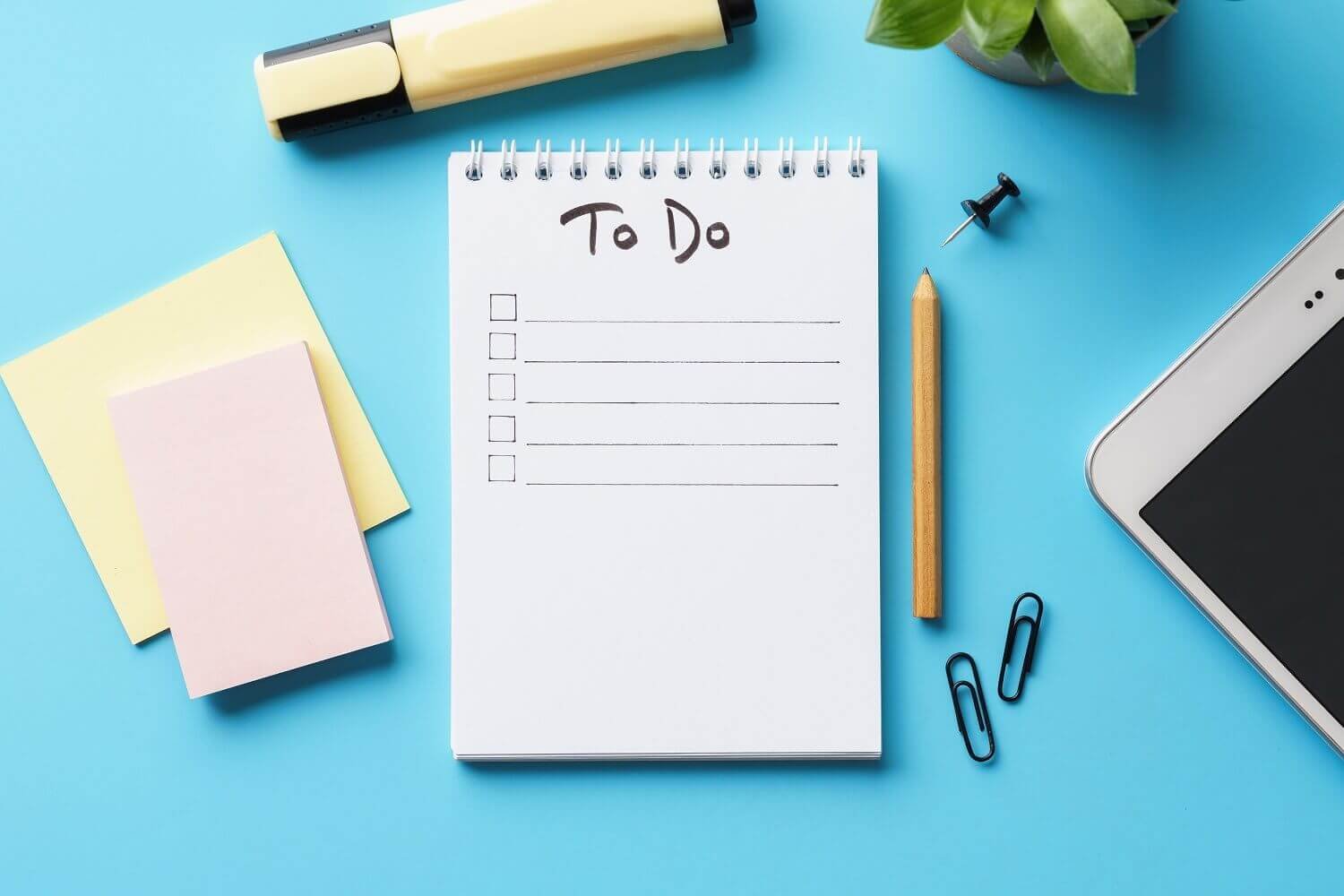A blue desk with a to-do list notepad, yellow sticky notes, a pencil, paper clips, a push pin, a highlighter, and a smartphone, with a potted plant in the top right corner.