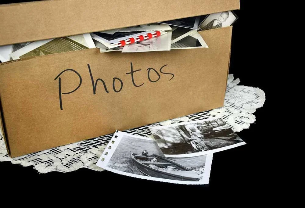 Brown cardboard box labeled 'Photos' with black marker, filled with printed black and white photographs, some scattered on a lace doily.