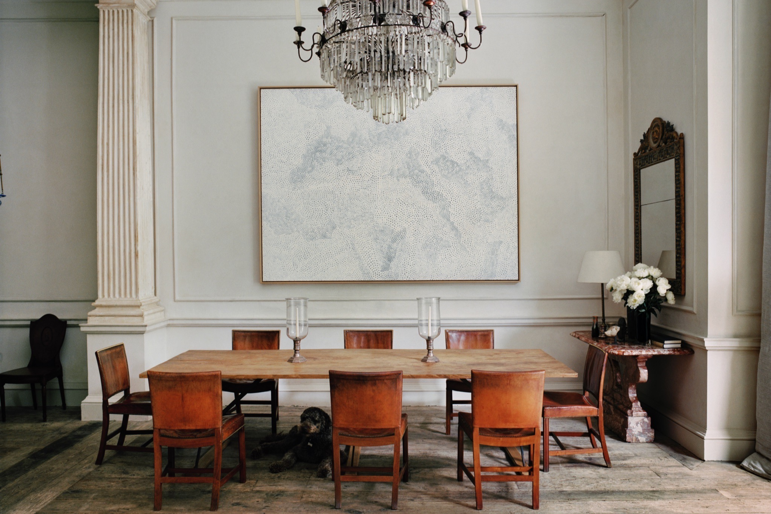 A dining room with a large wooden table, eight wooden chairs, and a black dog lying under the table. The room has light-colored walls, a large framed map of Europe on the wall, and a chandelier hanging from the ceiling. To the right, there is a side 
