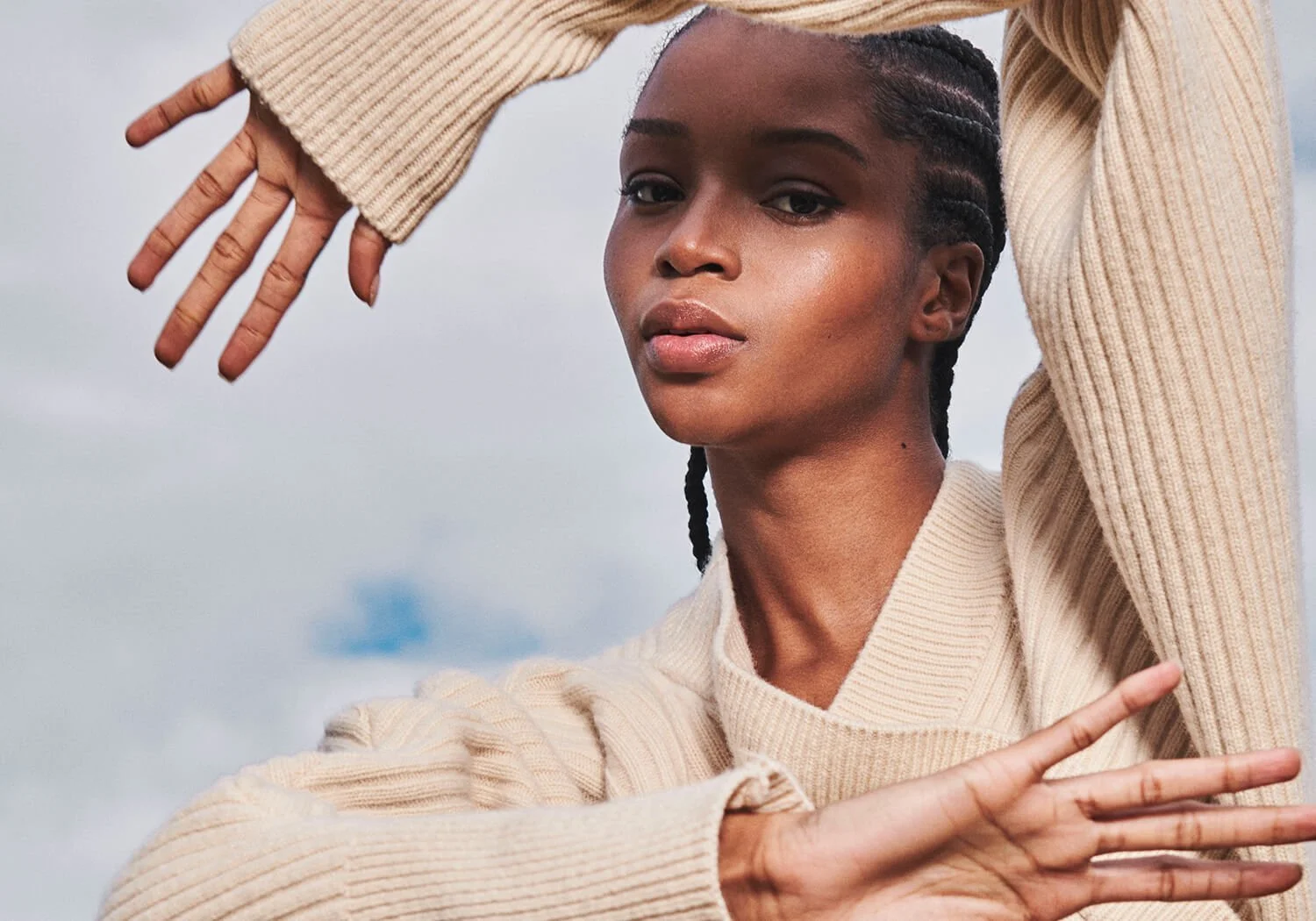A young woman with dark braided hair is posing outdoors against a cloudy sky background, with her arms extended and hands open in a dynamic pose, wearing a beige ribbed sweater.