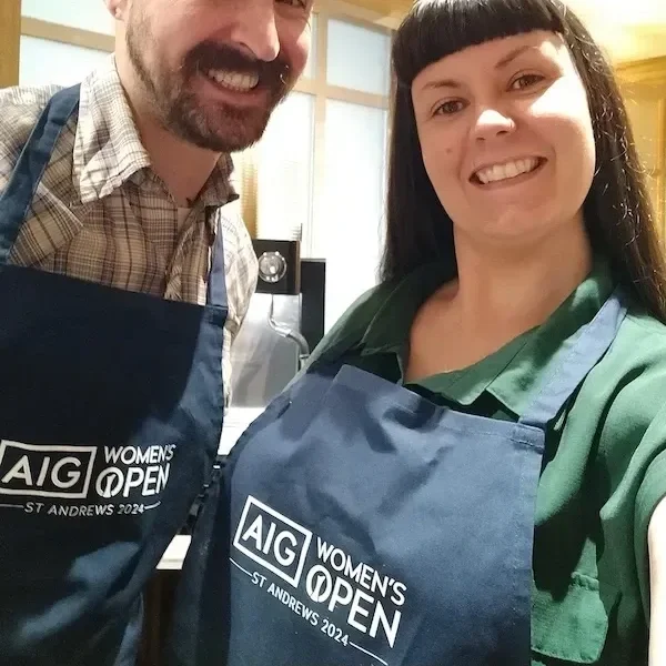 Two smiling women wearing blue aprons with 'AIG Women's Open, St. Andrews 2024' logo, in a kitchen or food prep area.