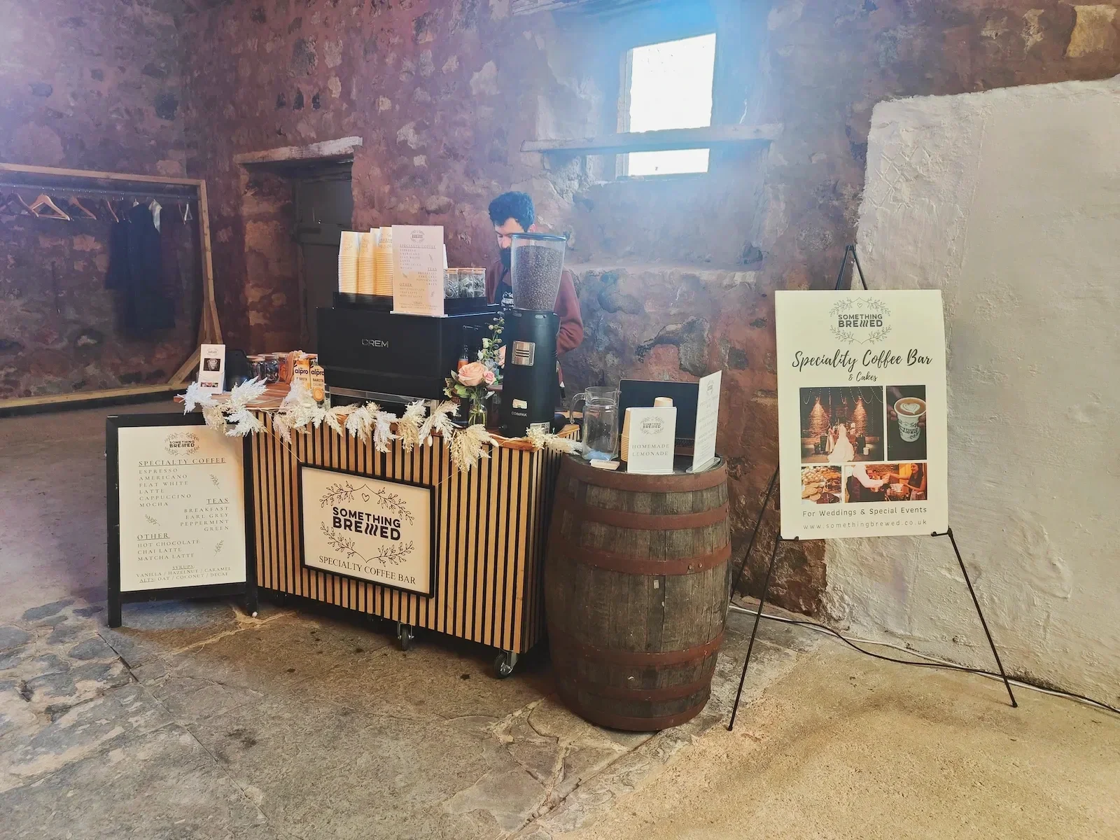 A coffee stand called 'Something Brewed' with a menu, decorated with white foliage and pink flowers, set inside a rustic brick-walled space. A barista is behind the counter, and a large sign on an easel promotes the specialty coffee bar for weddings and events.