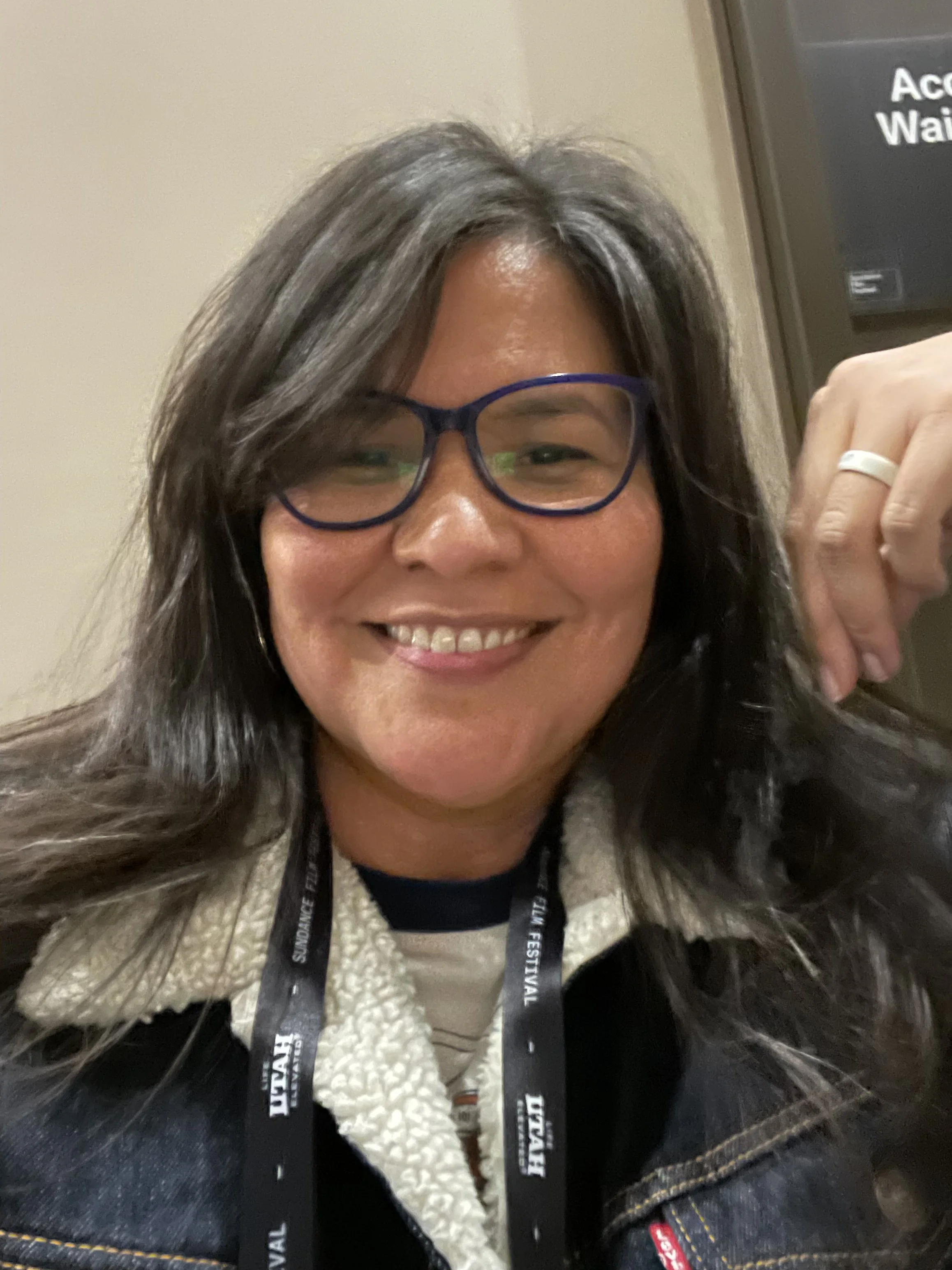 Smiling woman with long dark hair, glasses, and a denim jacket taking a selfie at an event, with a lanyard around her neck.