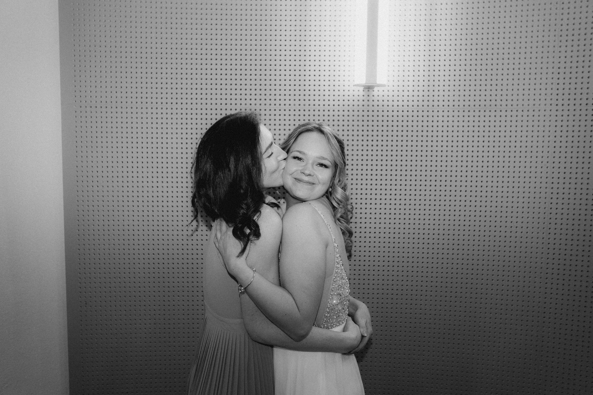 Zwei Frauen umarmen sich und küssen sich, eine lächelt in die Kamera auf einem perforierten Hintergrund in Schwarz-Weiß.