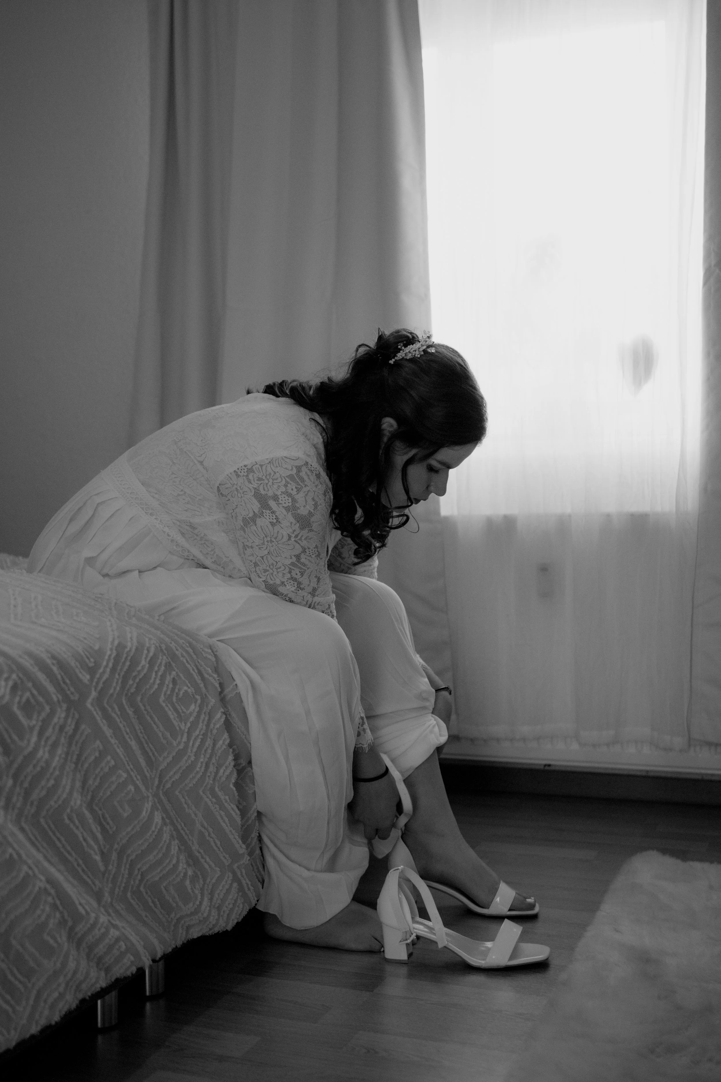 Frau in Hochzeitskleid sitzt auf dem Bett und zieht ihre Schuhe an, vor einem hell erleuchteten Fenster.