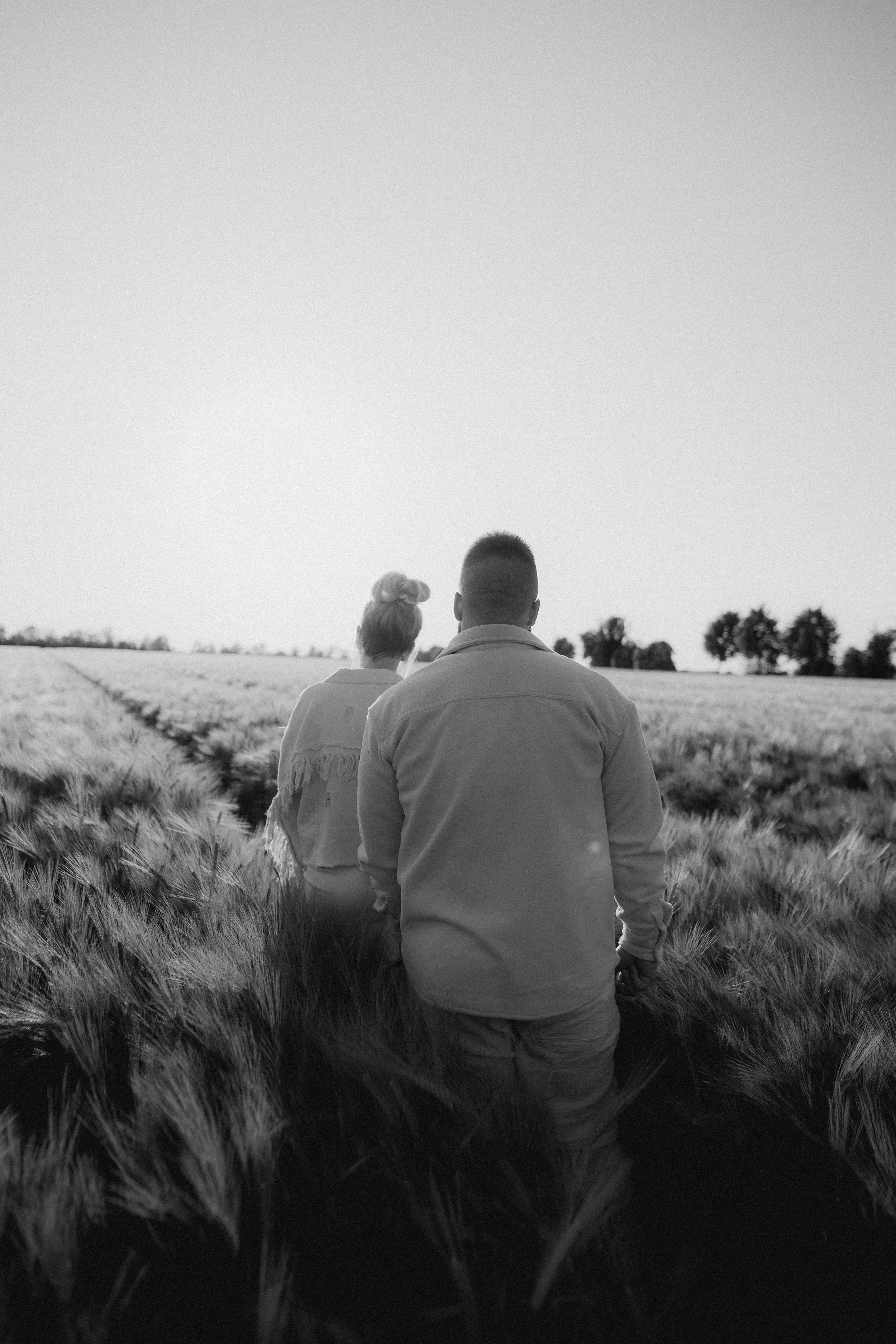 Zwei Menschen gehen durch ein Weizenfeld in Schwarz-Weiß, Blick nach hinten, im Hintergrund Bäume, hoher Himmel.
