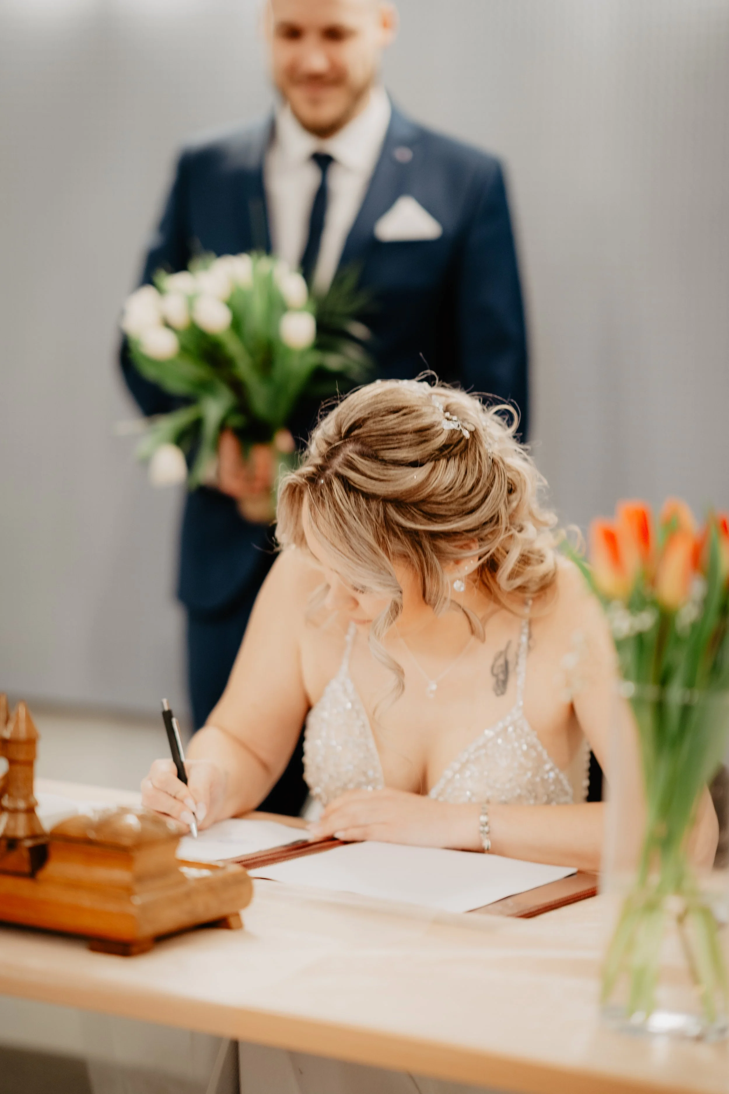 Eine Braut unterschreibt eine Urkunde bei ihrer Hochzeit, während ein Bräutigam im Hintergrund eine Blumenstrauß hält.