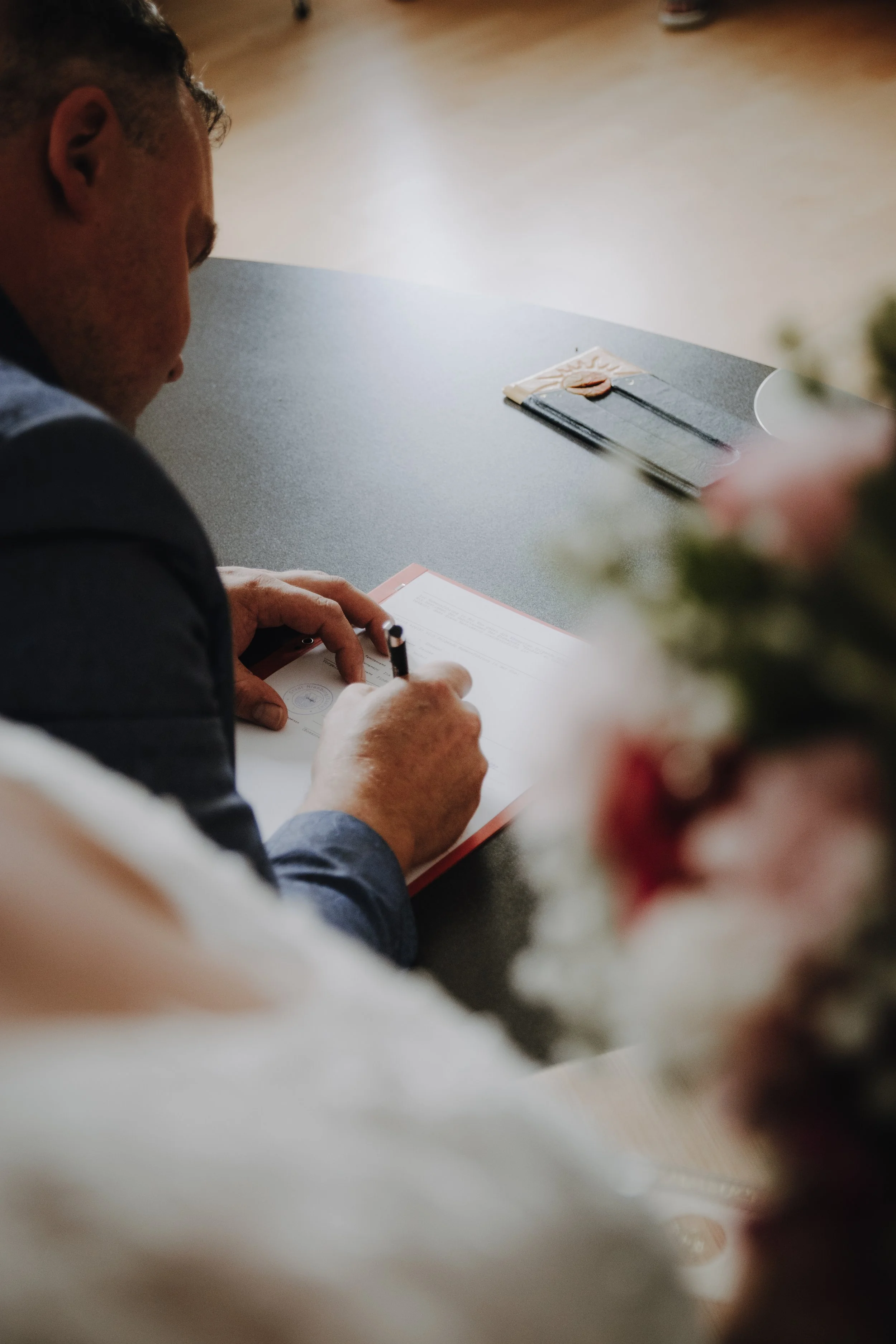 Ein Mann unterschreibt ein Dokument bei einer Zeremonie, wahrscheinlich eine Heirat. Im Vordergrund sind Blumen leicht unscharf.