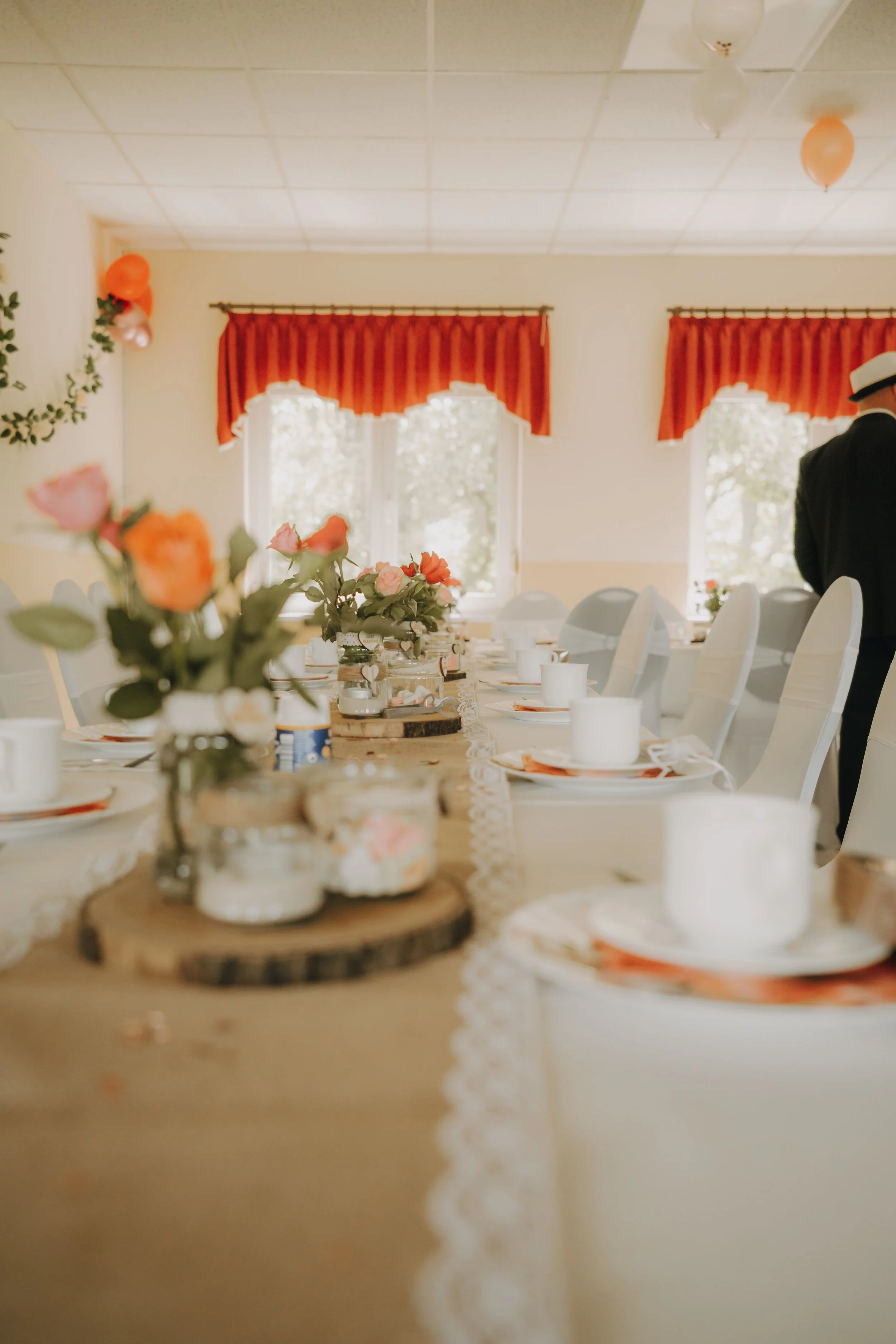 Ein festlich gedeckter Tisch mit Blumen, Kerzen und Tellern in einem Raum mit Fenster und roten Vorhängen, auf dem ein Mann in Anzug und Hut steht.