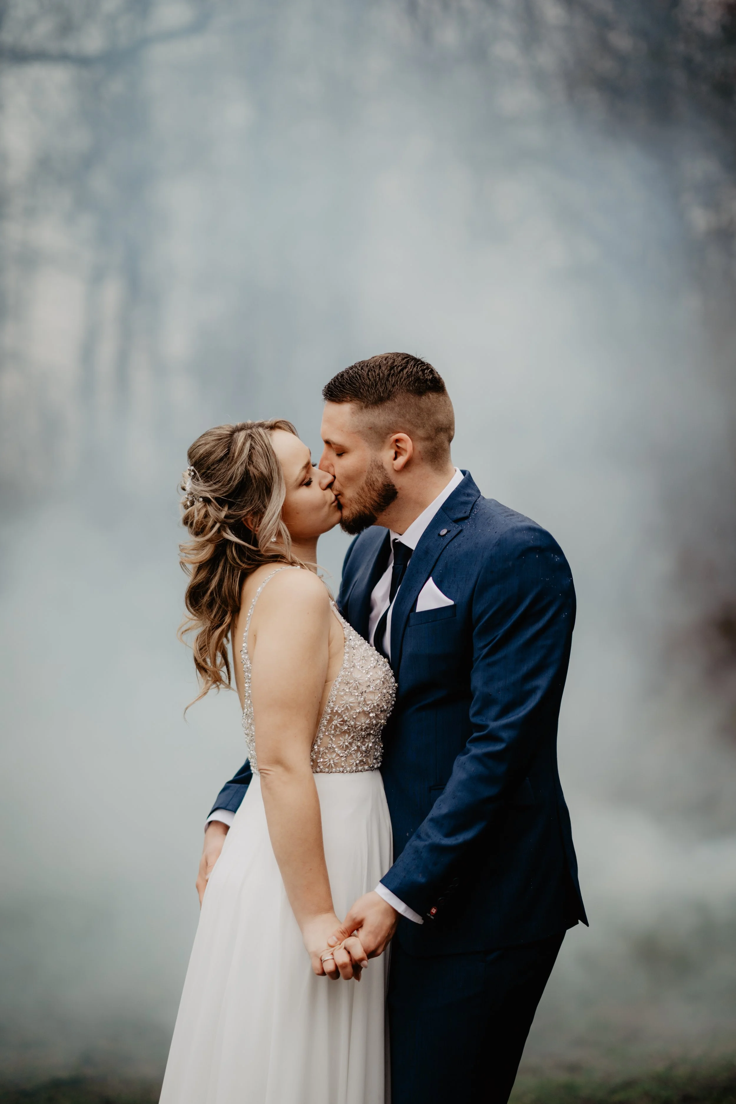 Ein Brautpaar teilt einen Kuss im Freien mit nebliger Hintergrundkulisse.