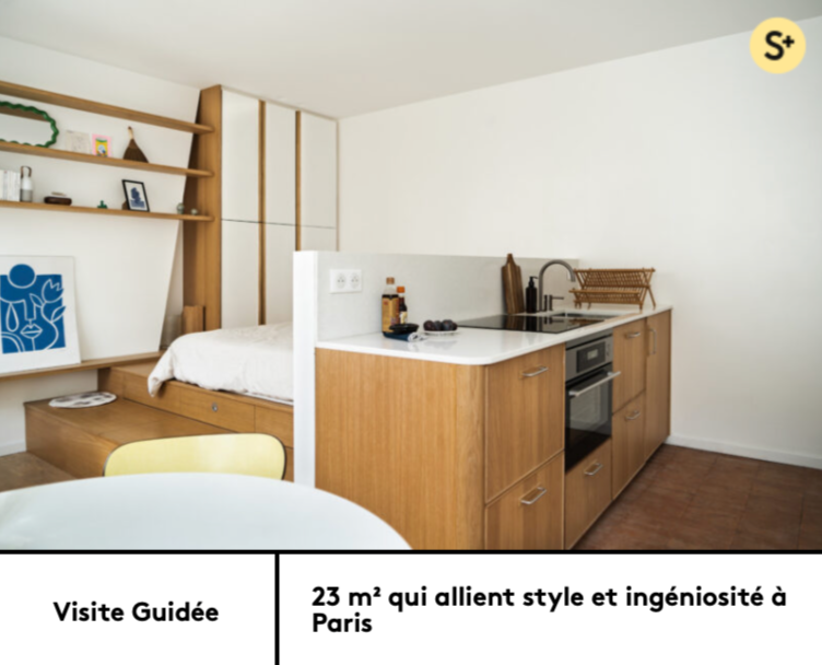 Coin cuisine avec comptoir en bois, plaque de cuisson électrique, étagères en bois, décor minimaliste, espace intégré dans un studio avec coin nuit visible en arrière-plan.
