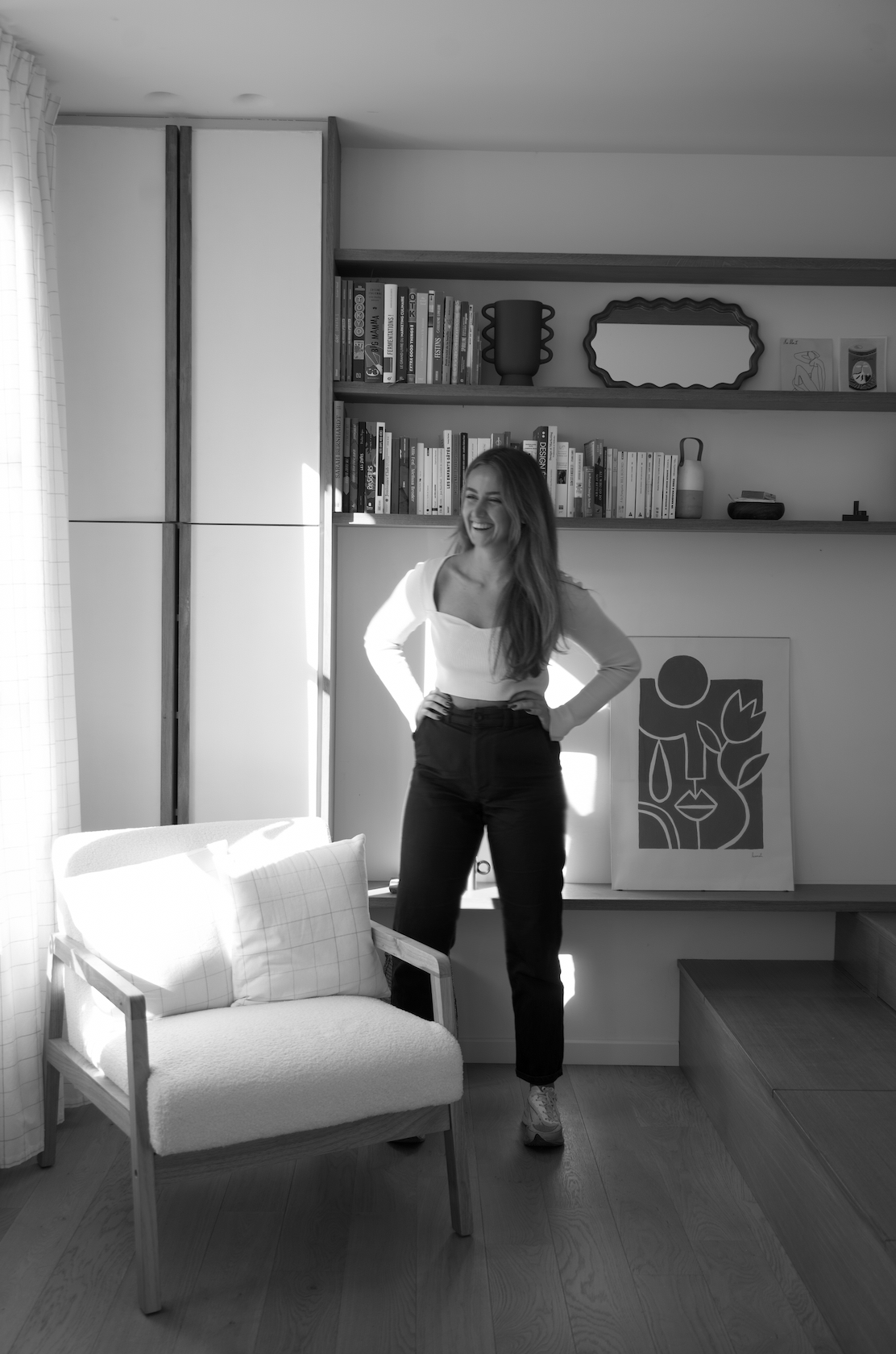 Une femme souriante se tenant dans un salon moderne en noir et blanc, à côté d'une bibliothèque et d'une œuvre d'art abstraite derrière elle.