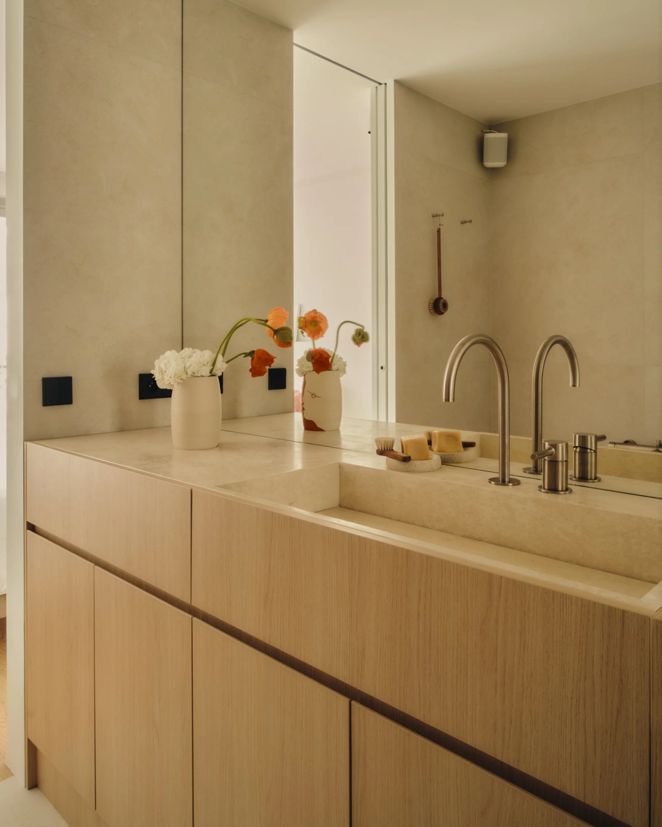 Intérieur d'une salle de bain avec un lavabo en pierre, deux vasques, robinets métalliques, deux vases avec des fleurs, et une décoration minimaliste en bois et teintes neutres.