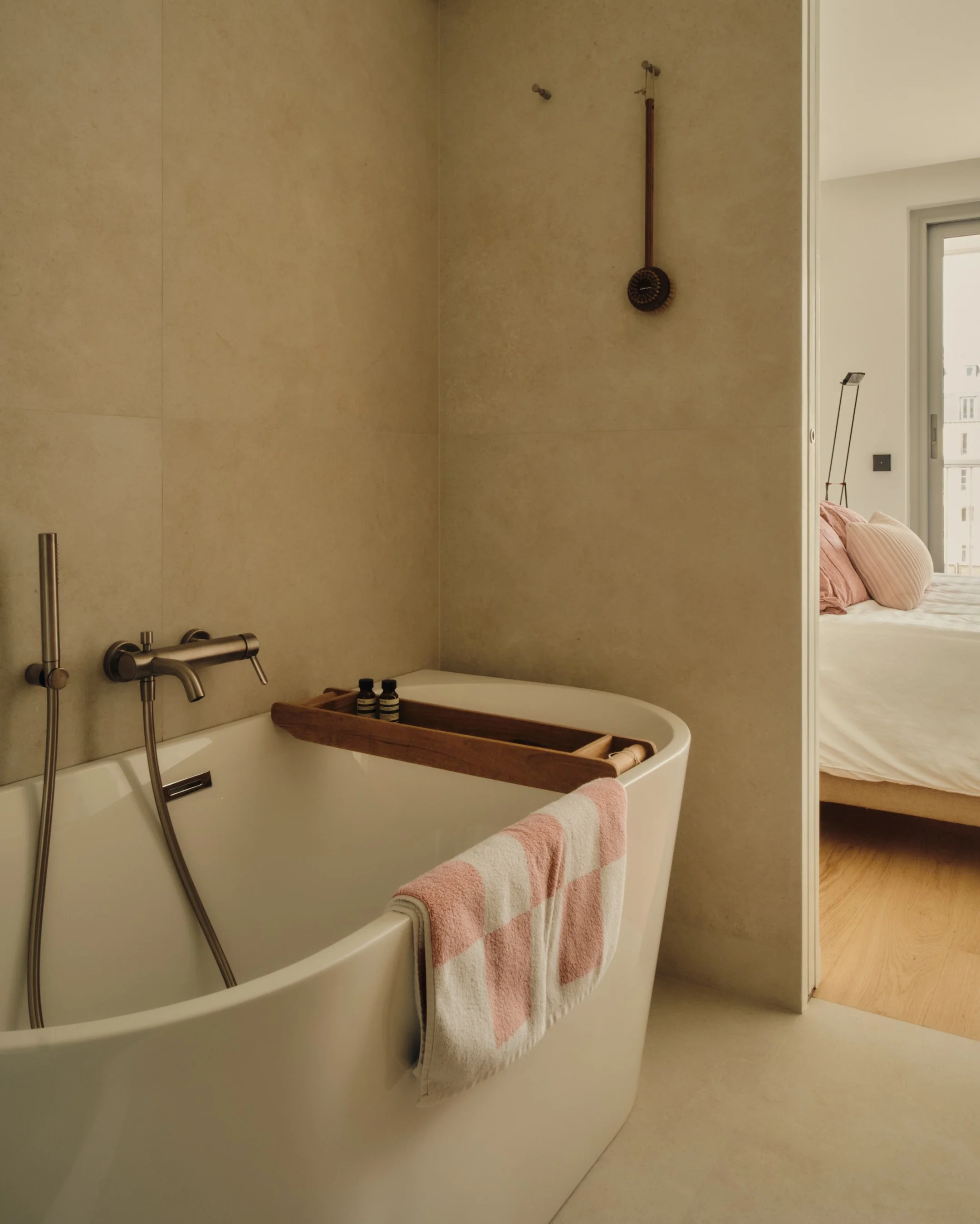 Baignoire blanche dans une salle de bain minimaliste avec une serviette rose et blanche suspendue à l'extérieur, éléments de bain en bois et un mur beige. Exit la douche, un objet étrange suspendu au plafond. Chambre avec lit avec oreillers roses vis