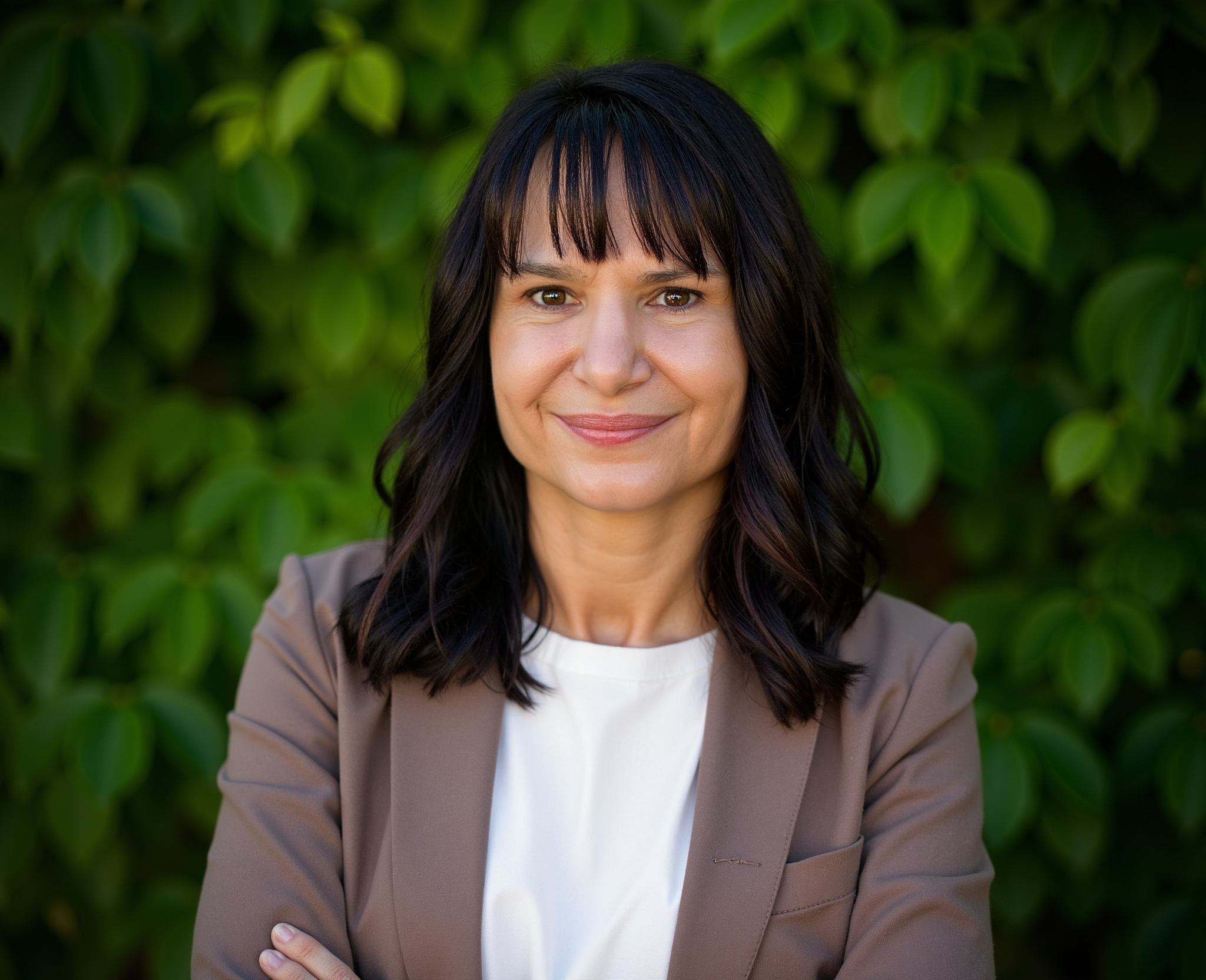 A woman with black hair and bangs smiling, standing outdoors with green leaves in the background.