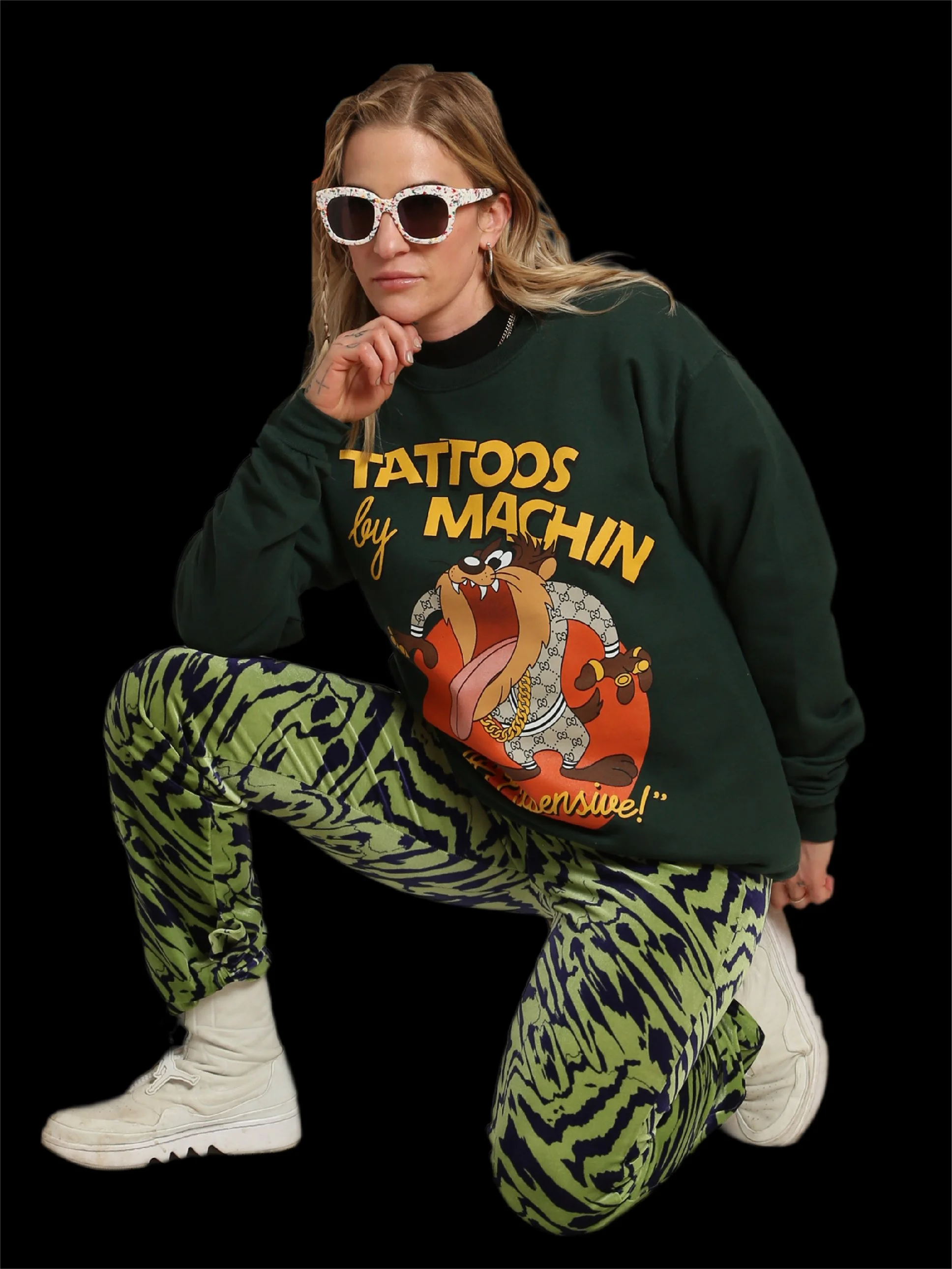 Woman wearing white sunglasses, a green sweatshirt with cartoon character and text, patterned green and black pants, and white sneakers, kneeling in a pose against a black background.
