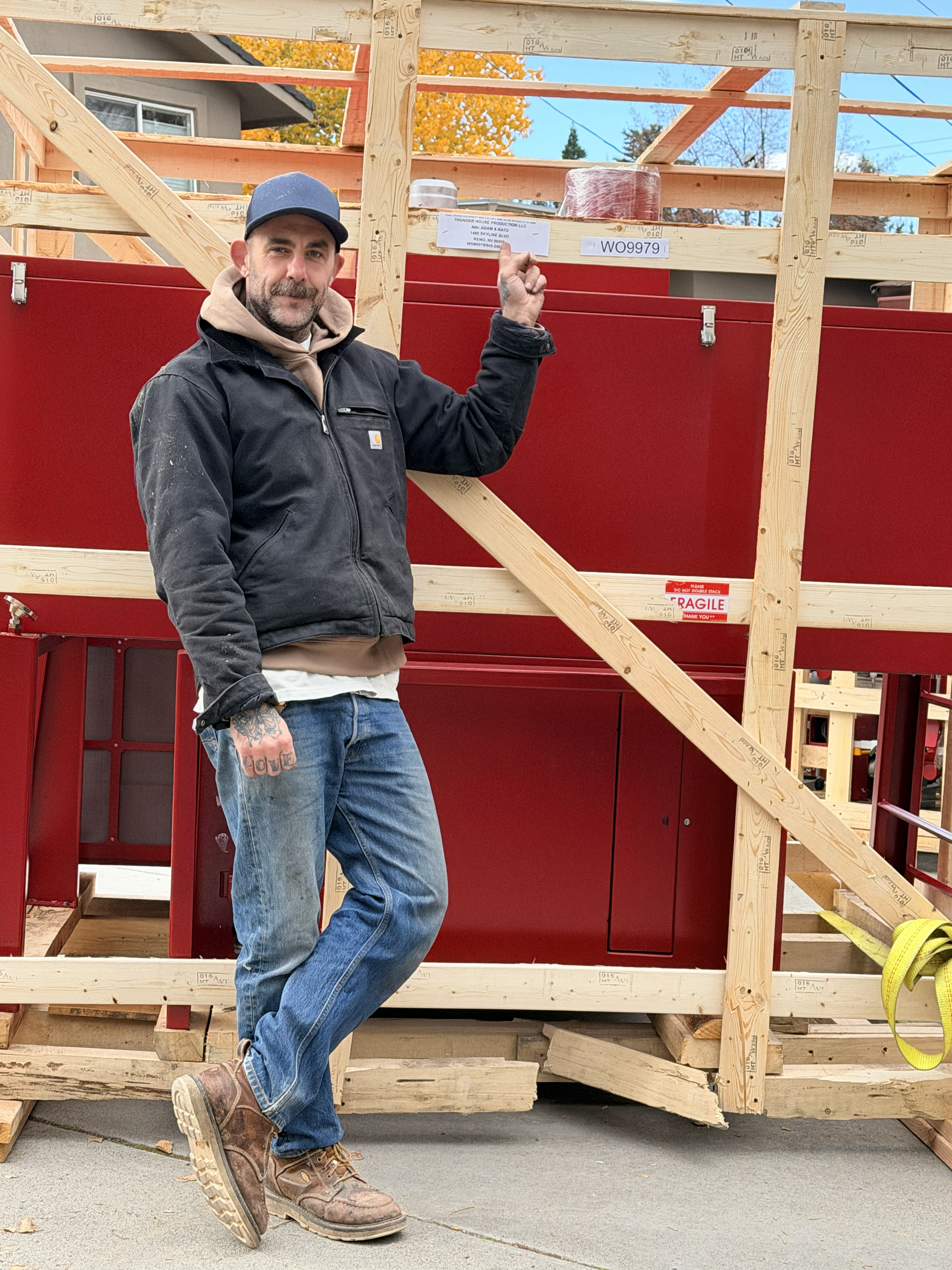 A man standing on a construction site in front of a partly built wooden structure, pointing at a label on the framework, with a red cabinet and construction materials around him.