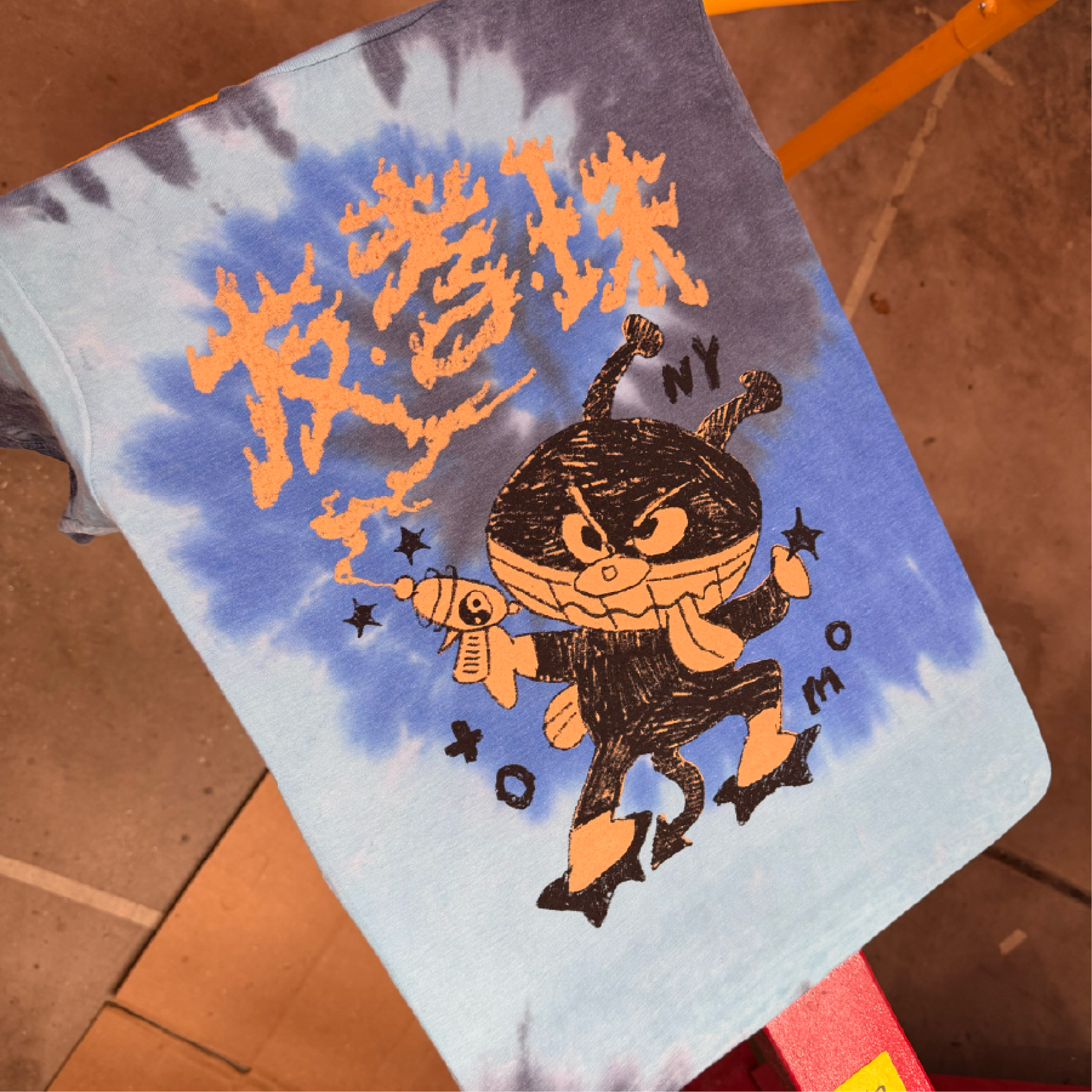 A close-up of a baseball cap with orange and black graphics, including a pumpkin face, a spider web pattern, and text reading 'Halloween NY.' The cap is resting on a brown tiled floor with an orange table or barrier in the background.