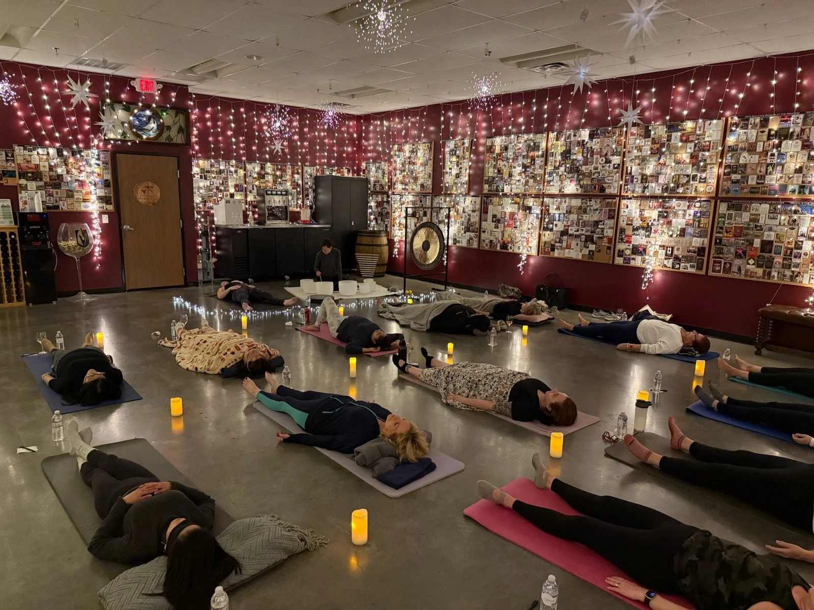 People participating in a group yoga class in a decorated studio, with string lights, hanging paper stars, and a collage of pictures on the wall, all illuminated by candles along the floor.