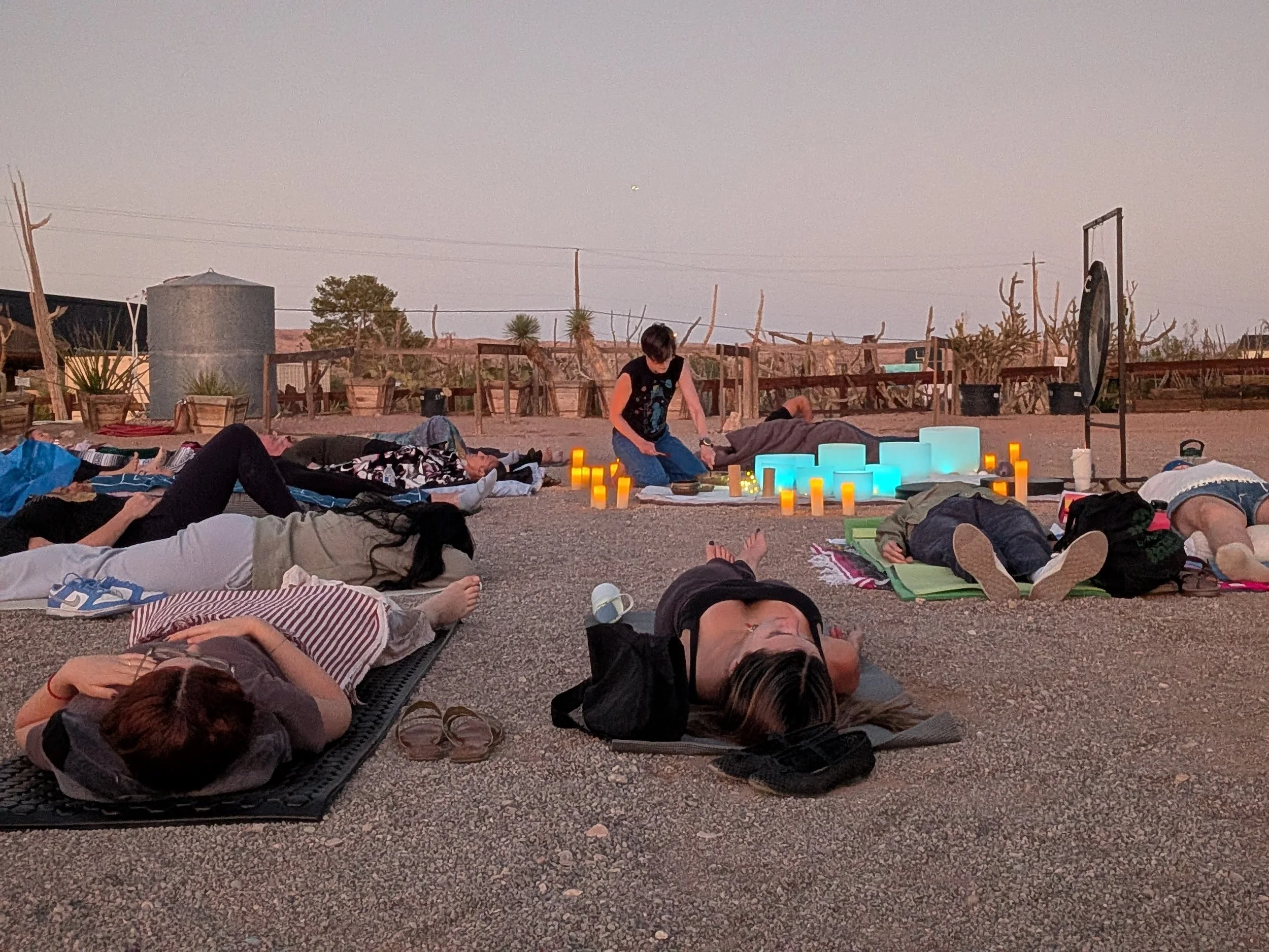 People lying on yoga mats and blankets outdoors at dusk, participating in a group meditation or yoga session with glowing blue and yellow candles and lanterns, desert plants, and a water tank in the background.