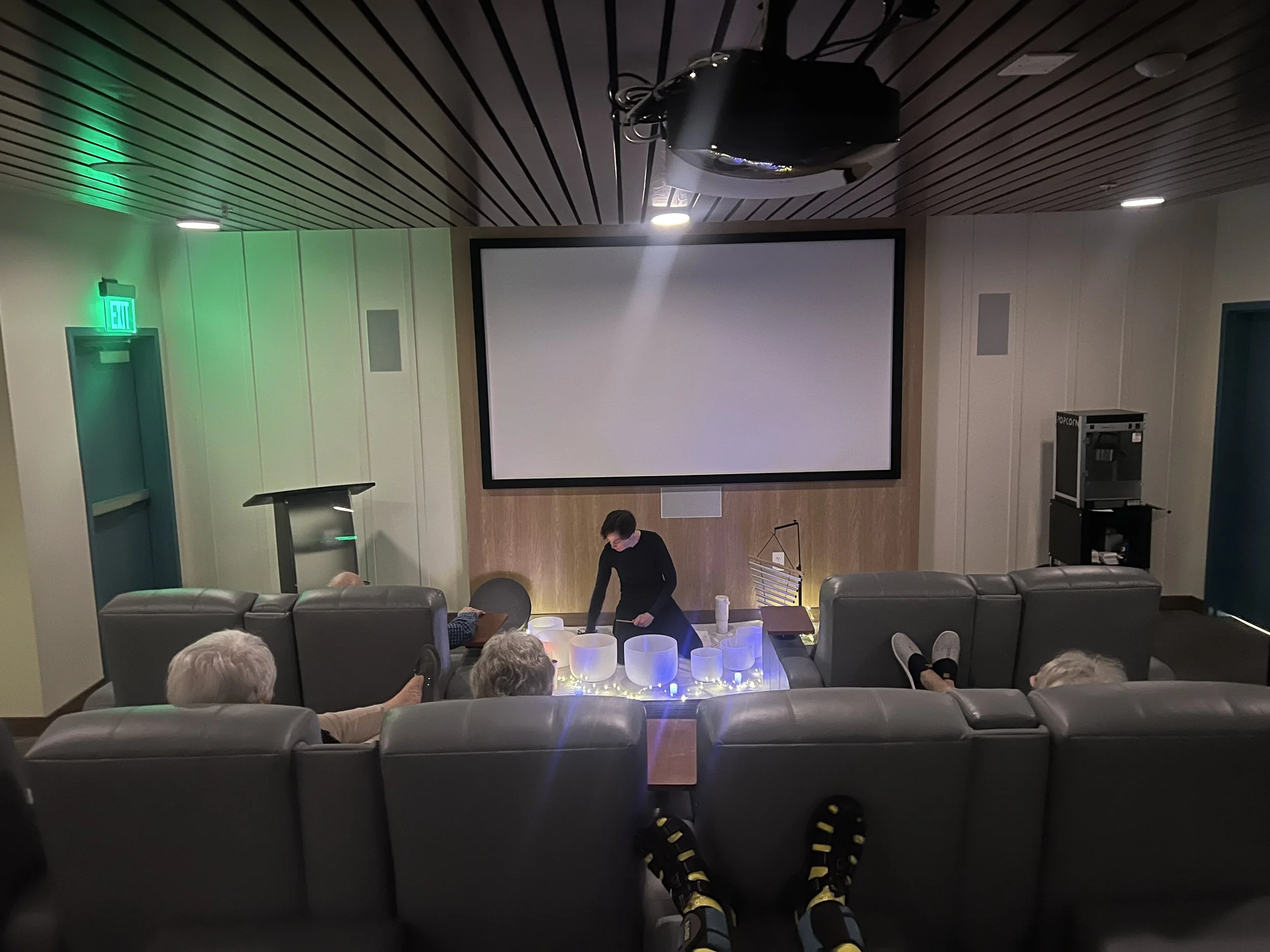 Small theater or screening room with a large blank projection screen, four elderly people sitting on dark gray couches, and a woman standing in front of decorative lighting on the floor. The ceiling is wooden with recessed lighting, and there is a popcorn machine in the back corner.