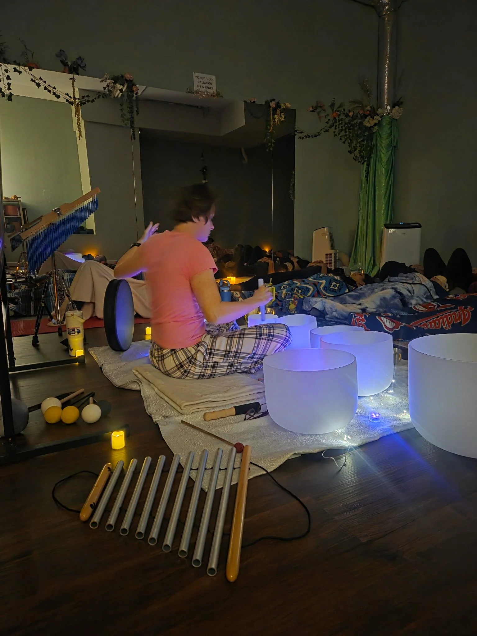 A woman sitting on a blanket playing crystal singing bowls, surrounded by other instruments and people lying down, in a dimly lit room decorated with flowers, fairy lights, and green curtains.