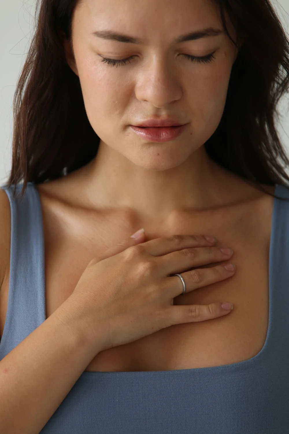 A woman with closed eyes and dark hair holds her hand over her chest, wearing a blue sleeveless top and a ring on her finger.
