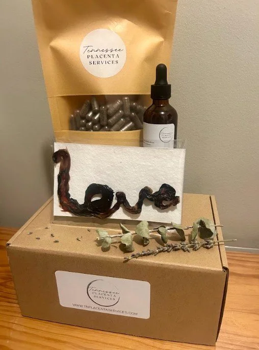 A placenta encapsulation package, cardboard box and a paper bag containing herbal products, a placenta tincture, a dried plant, and a card with text, arranged on a wooden table.