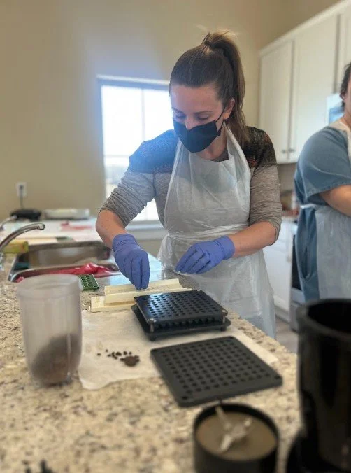 A woman wearing a face mask, gloves, and apron working with a small object in a kitchen, preparing placenta for encapsulation