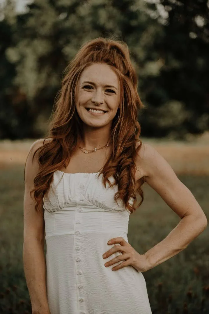 A smiling woman with long, wavy red hair wearing a white strapless dress and jewelry, standing outdoors on a grassy area with trees in the background during daylight.