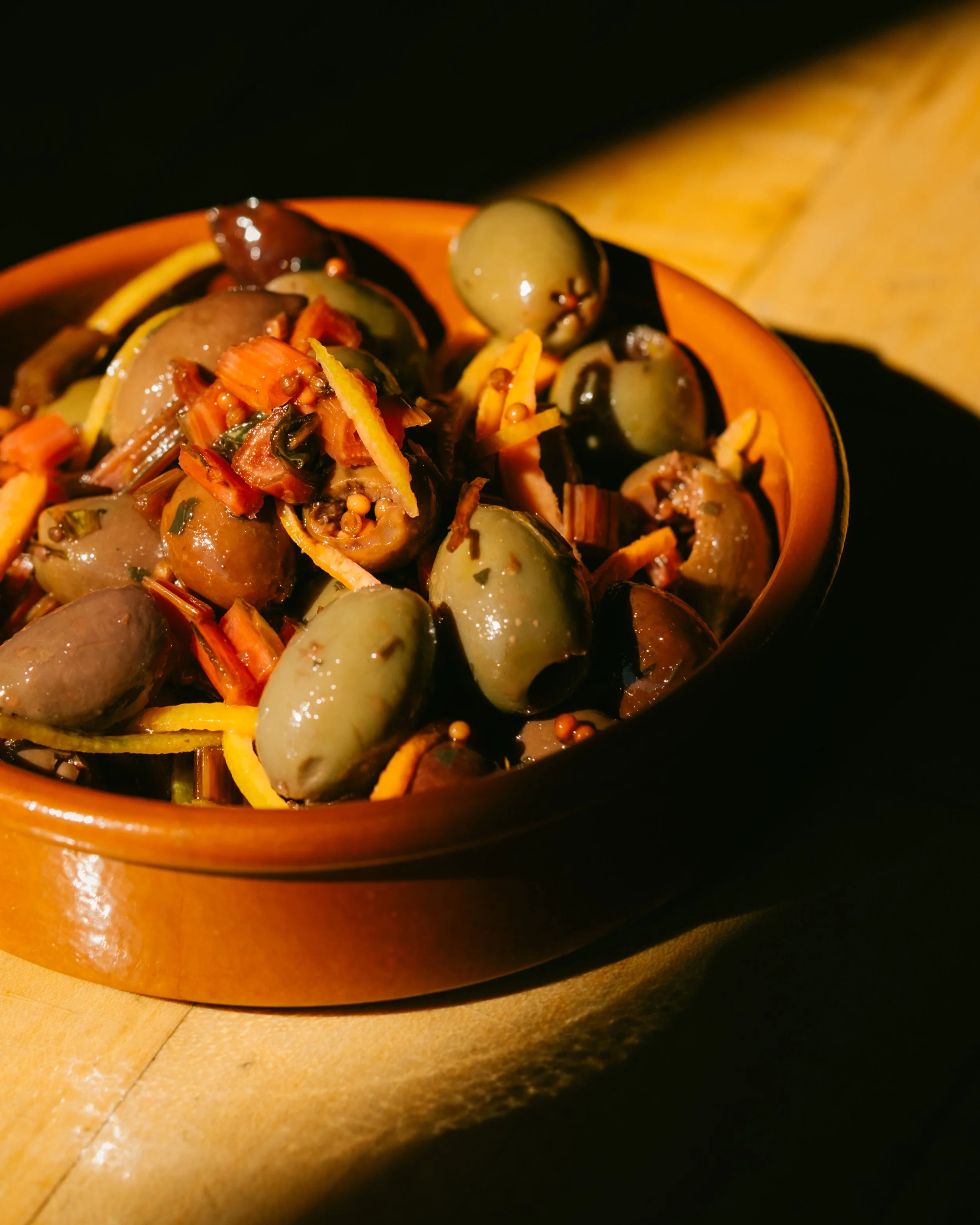 Close-up of a ceramic bowl filled with green grapes, sliced carrots, and possibly pickles or other vegetables, on a wooden table with a yellow cloth.