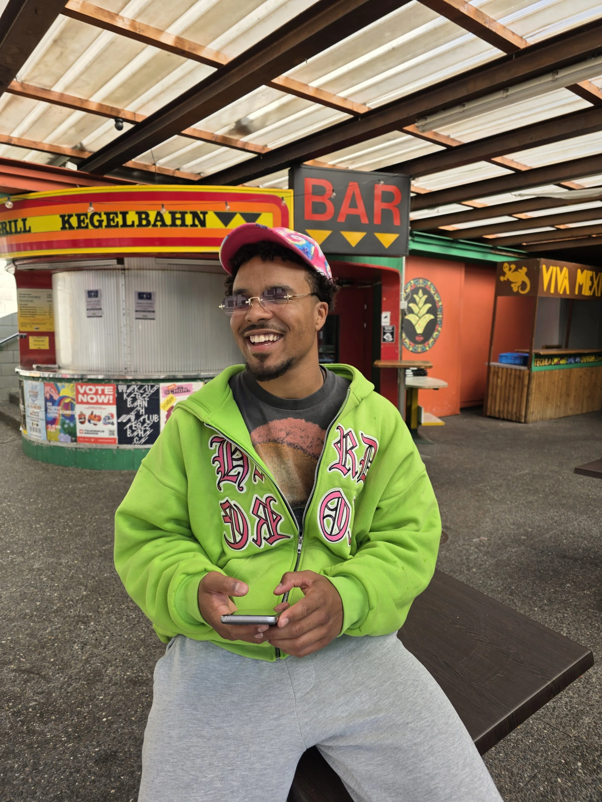 Ein lachender Mann in einer grünen Kapuzenjacke und grauen Hosen sitzt an einem Holztisch im Freien. Im Hintergrund sind Imbissstände mit Schildern wie 'Bar', 'Kegelbahn' und 'Viva Mexico' zu sehen.