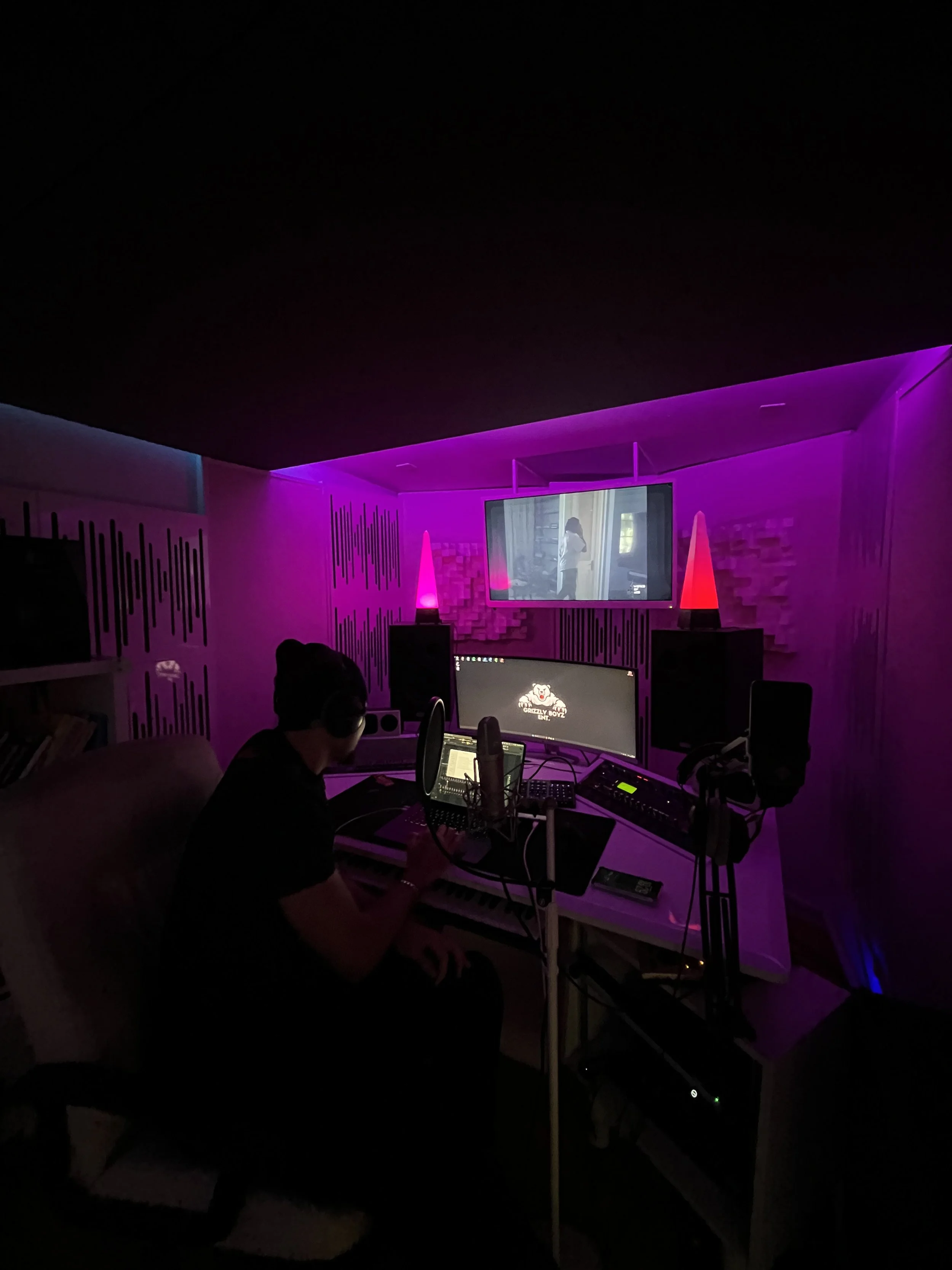 Ein Mann arbeitet in einem Tonstudio mit mehreren Monitoren, Mikrofonen und Audio-Equipment, das Raum ist in lila und pink beleuchtet.