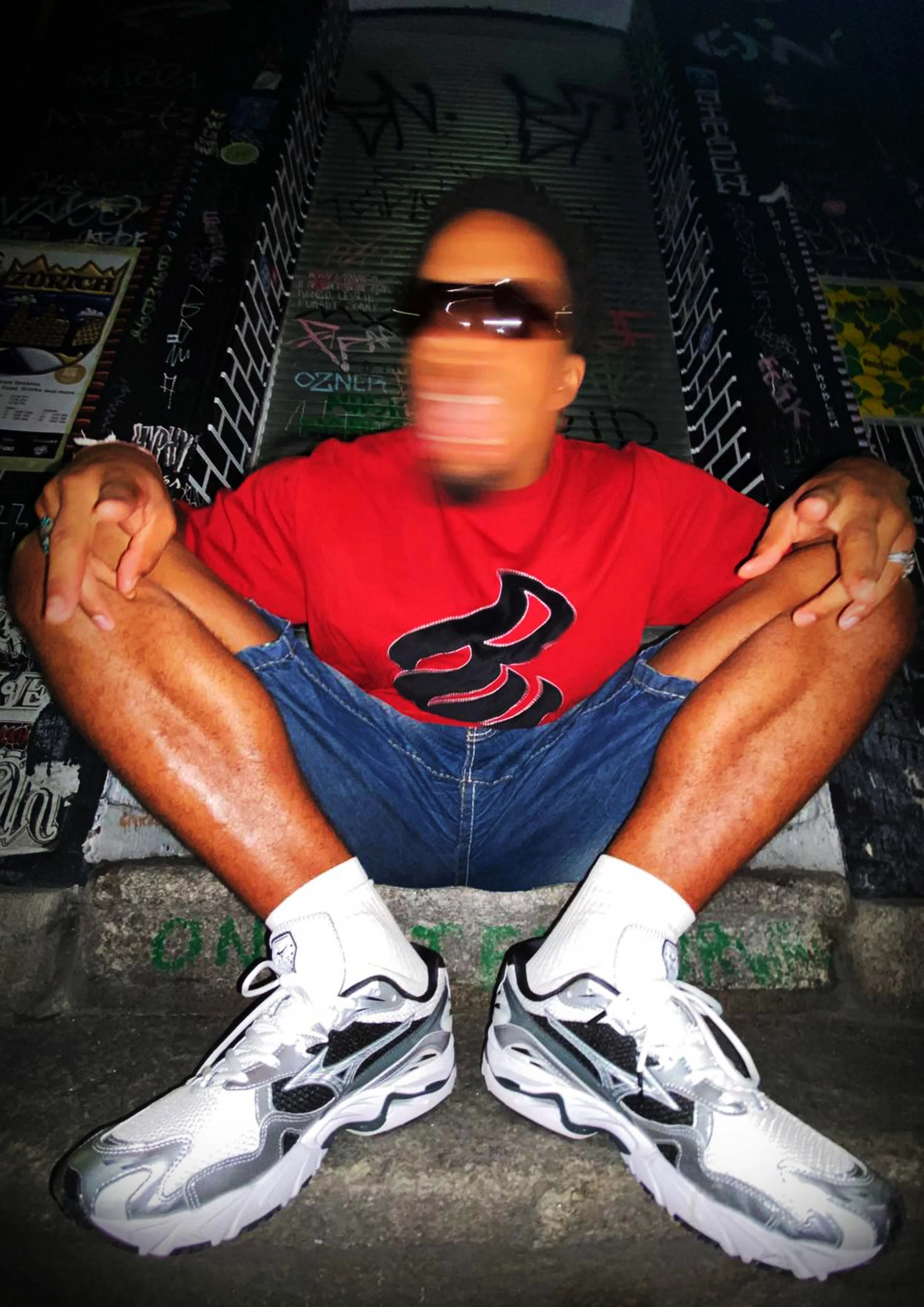 Junger Mann sitzend auf einer Treppenstufe, trägt weiße Sportschuhe, Jeansshorts, rotes T-Shirt, dunkle Sonnenbrille, mit verschwommenem Gesicht. Graffiti und Plakate im Hintergrund.