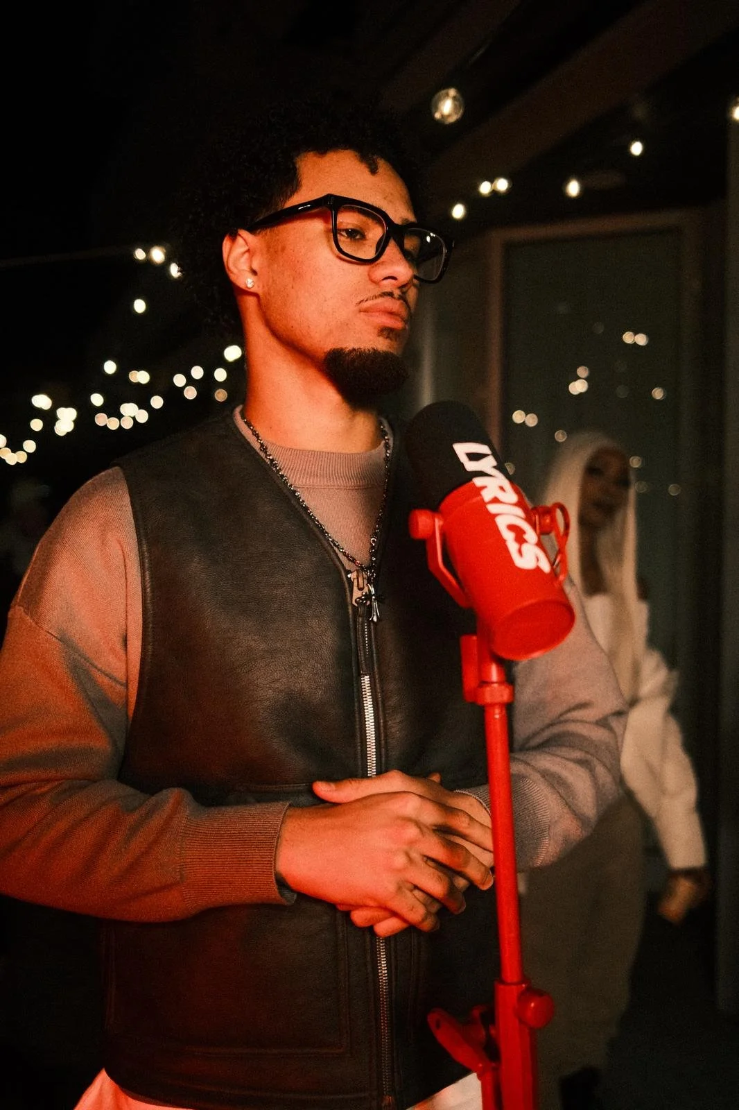 Ein junger Mann mit dunklem Afro, Brille und Bart steht vor einem Mikrofon bei einer Aufnahme im Aufnahmestudio, im Hintergrund eine blonde Frau.