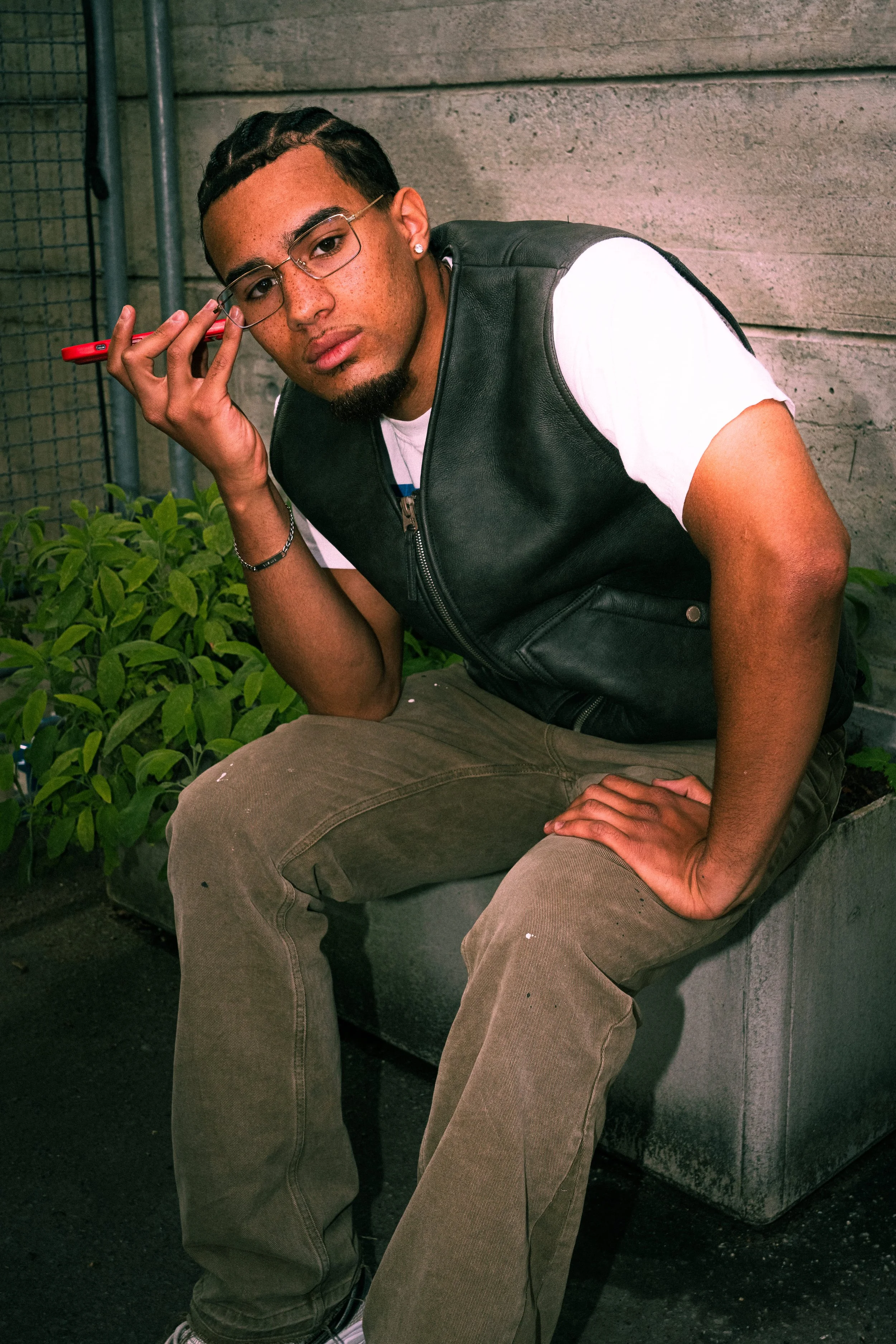 Junger Mann mit Brille und dunklen, gewellten Haaren, sitzend auf einer Betonkonstruktion, trägt eine schwarze Lederweste und helle Hosen, hält ein rotes Handy an sein Ohr, blickt in die Kamera.