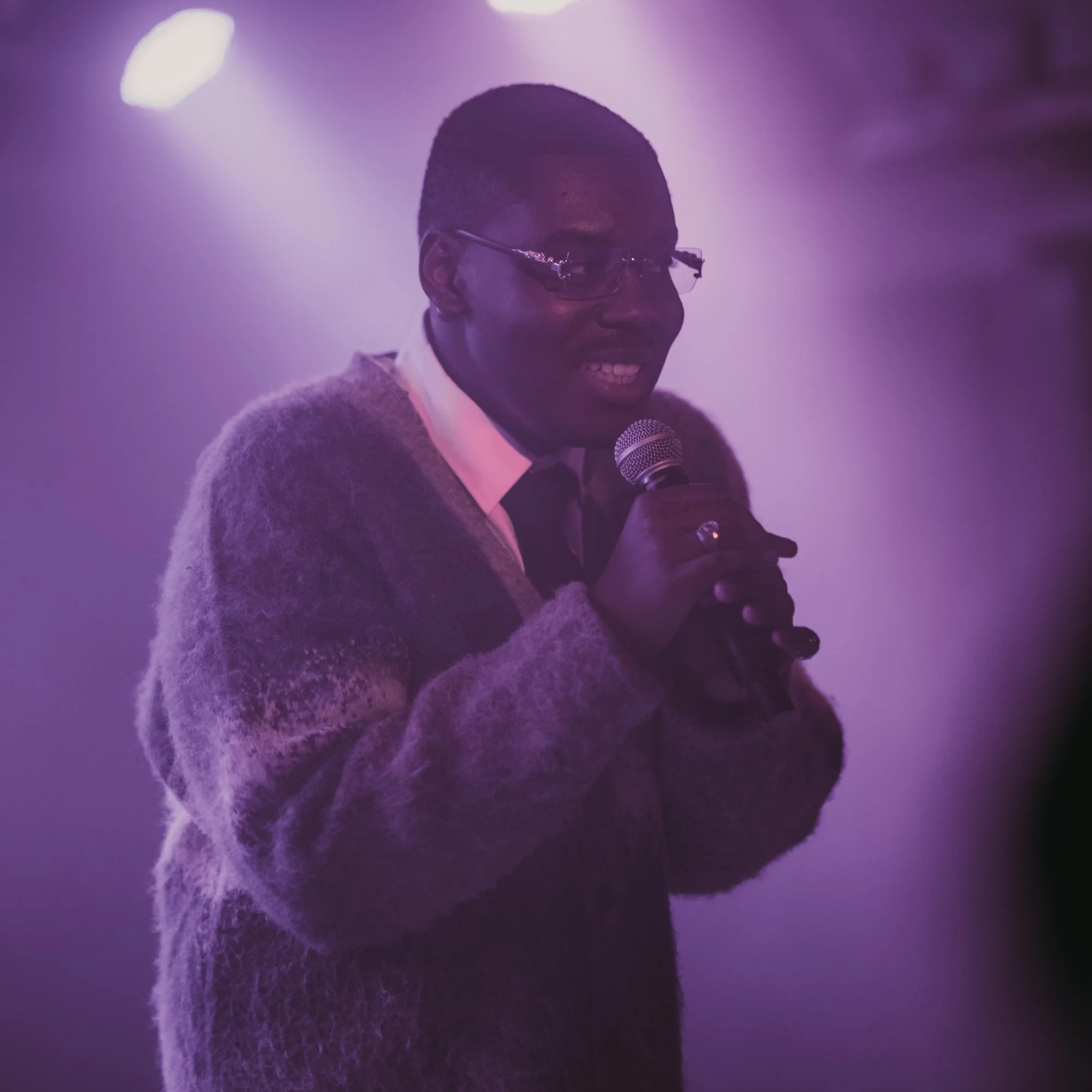 Eine Person mit dunkler Hautfarbe, die ein Mikrofon hält, während sie spricht, bei schwachem Licht mit einem violetten Farbton im Hintergrund.