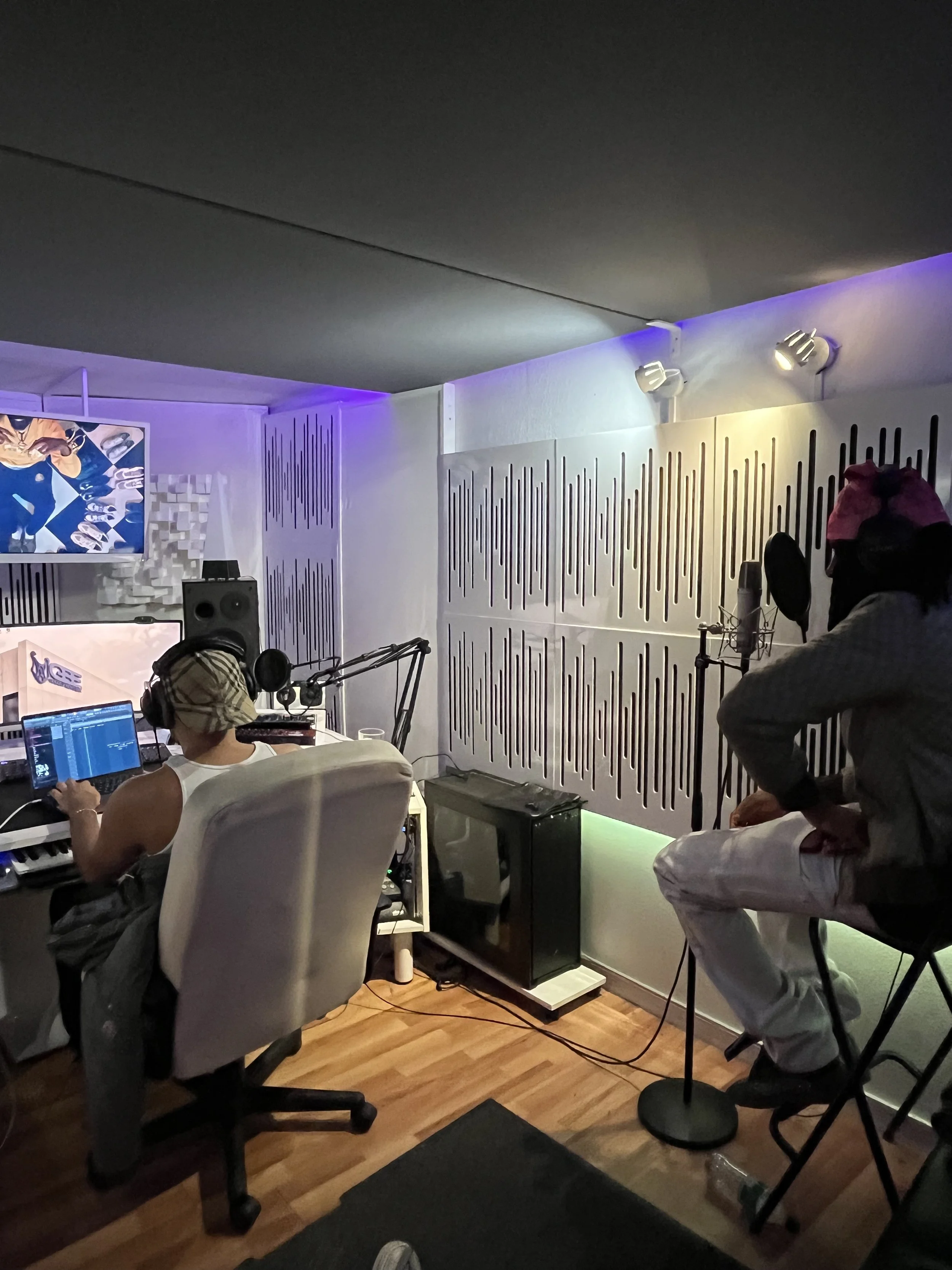 Zwei Personen in einem Tonstudio, einer arbeitet an einem Computer und der andere sitzt auf einem Stuhl mit einem Mikrofon, im Hintergrund sind schallabsorbierende Wandpaneele und eine LED-Beleuchtung.