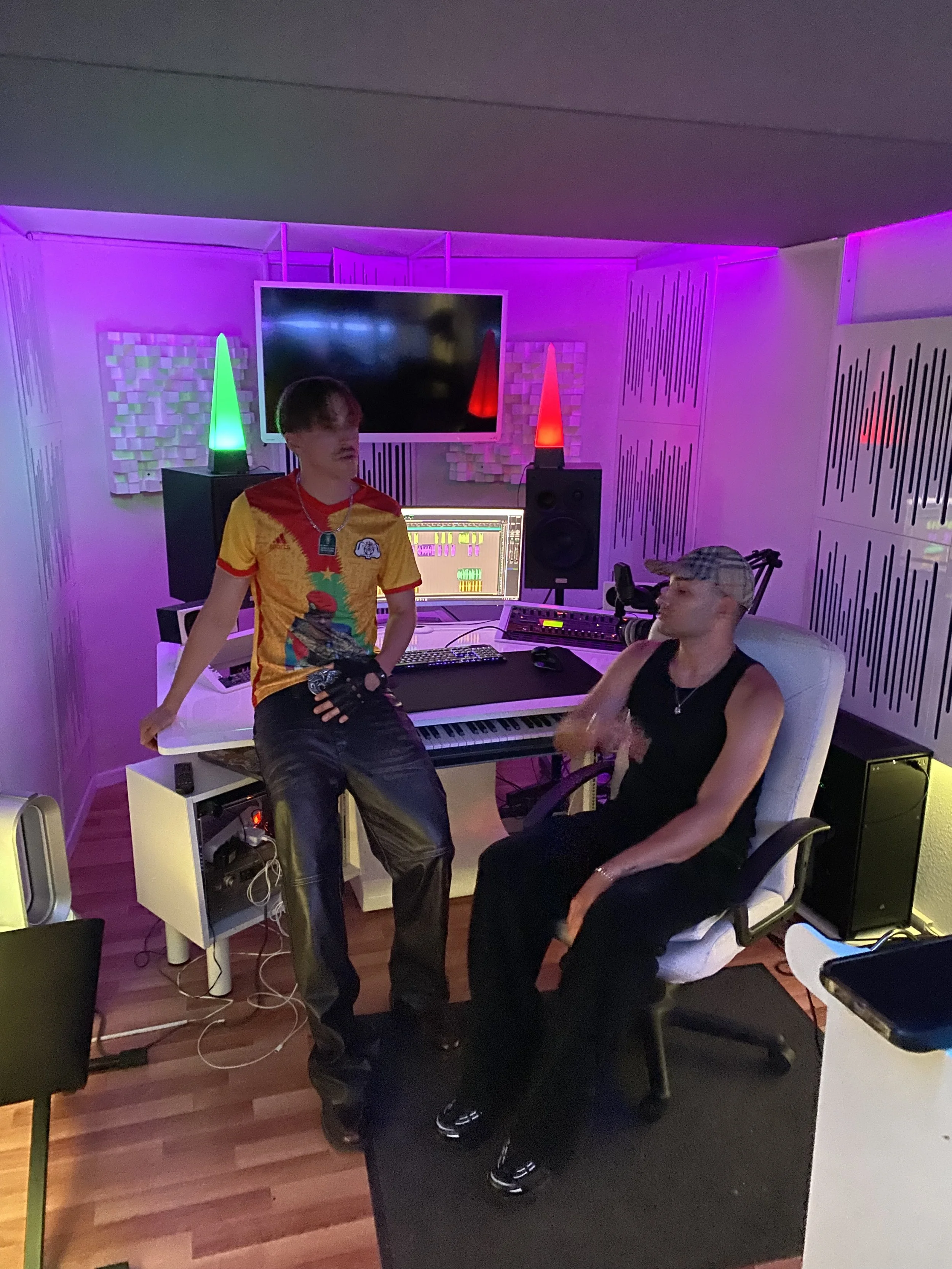 Zwei Menschen in einem Tonstudio, einer steht und der andere sitzt auf einem Schreibtisch, umgeben von Audioequipments, mit bunten Lichteffekten
