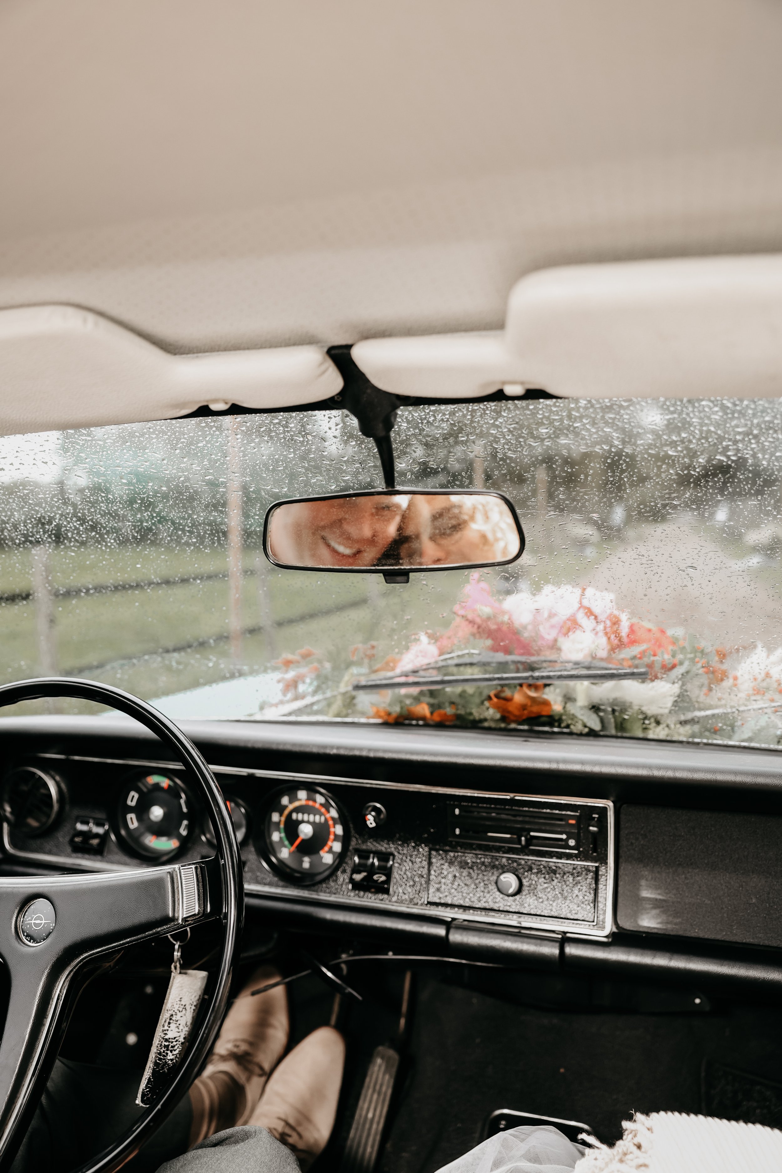 Binnenkant van een auto tijdens regen, met een achteruitkijkspiegel die reflecteert op een lachend paar mensen die achterin zitten, met bloemen op de voorruit.