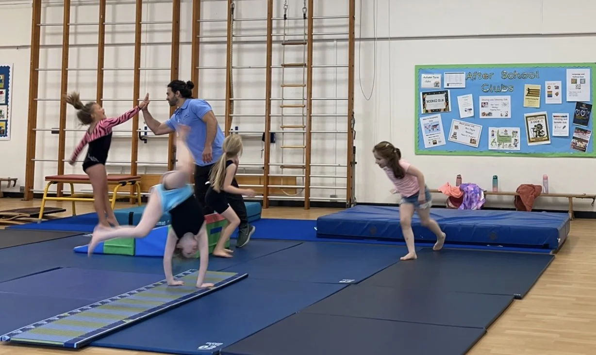 Children playing and practicing gymnastics on mats in a gymnasium with a teacher giving high-fives. There are gymnastic equipment and a bulletin board on the wall.