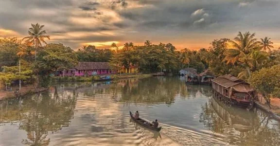 Vista de un río con casas en la orilla y un bote con una persona remando, en un atardecer con cielo nublado y árboles tropicales.