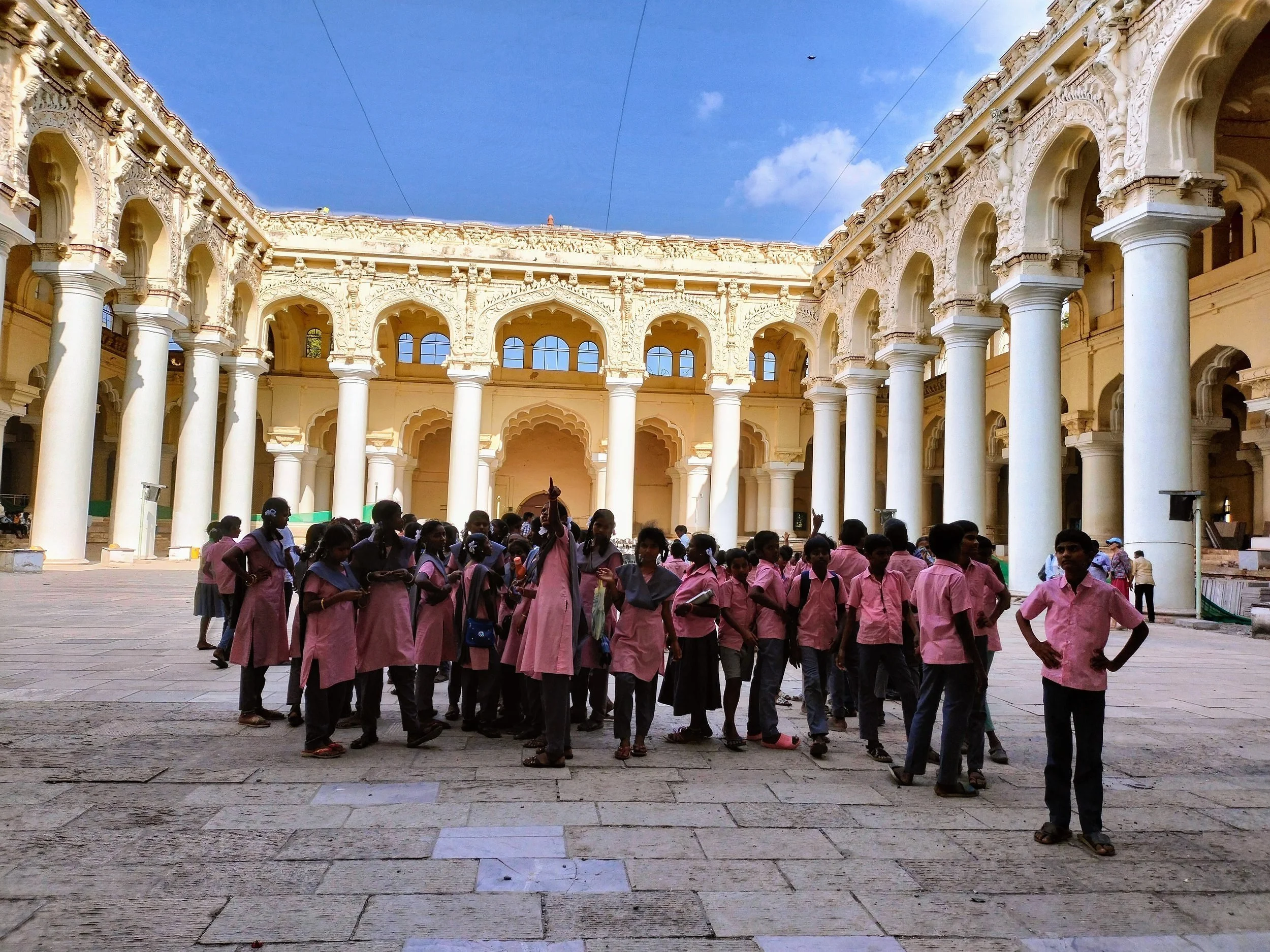 Grupo de niños en uniforme rosa reunidos en un atrio de arquitectura colonial con columnas blancas y detalles ornamentales, bajo un cielo azul con algunas nubes.